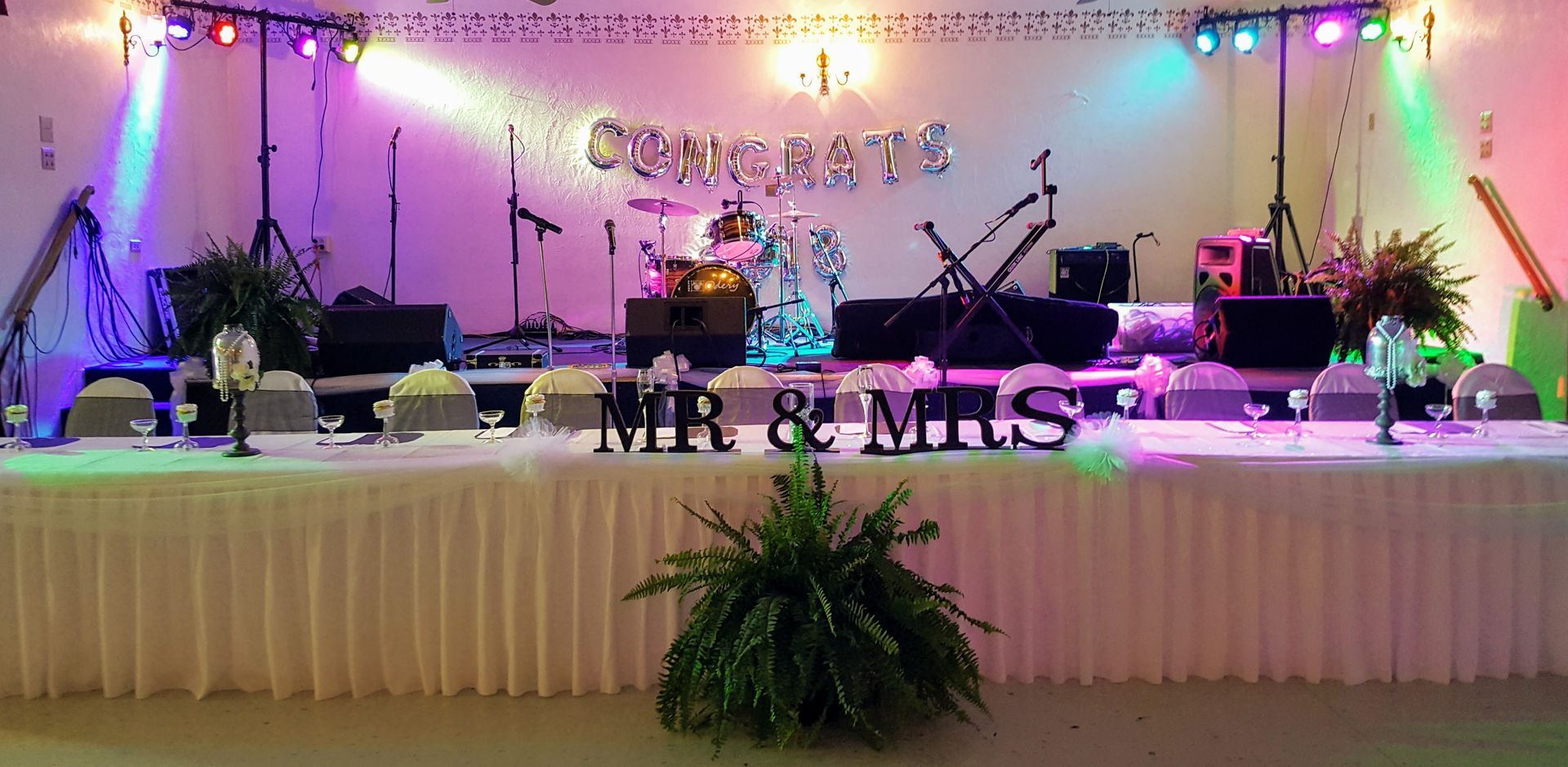 Decorated wedding reception stage with “MR & MRS” sign, fairy lights, floral accents, and microphones on both sides