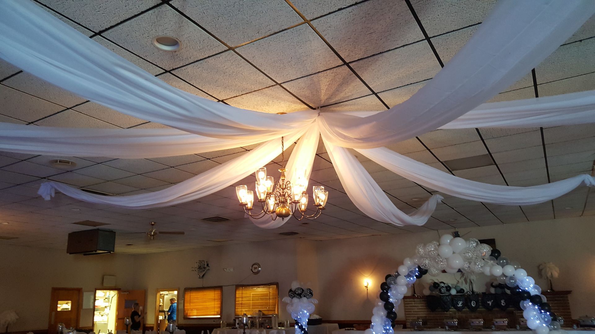 Decorated banquet hall ceiling with white drapes and a lit chandelier over tables and an arch of balloons