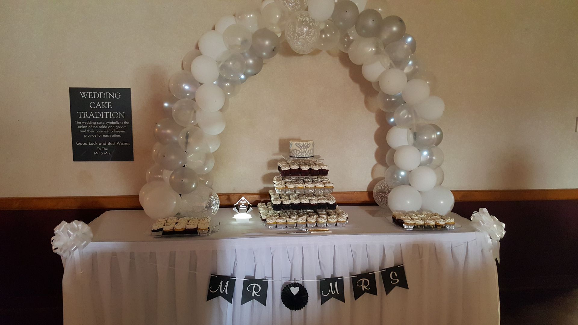 White balloon arch and tiered cake on a draped table with black-and-white bunting at an event backdrop