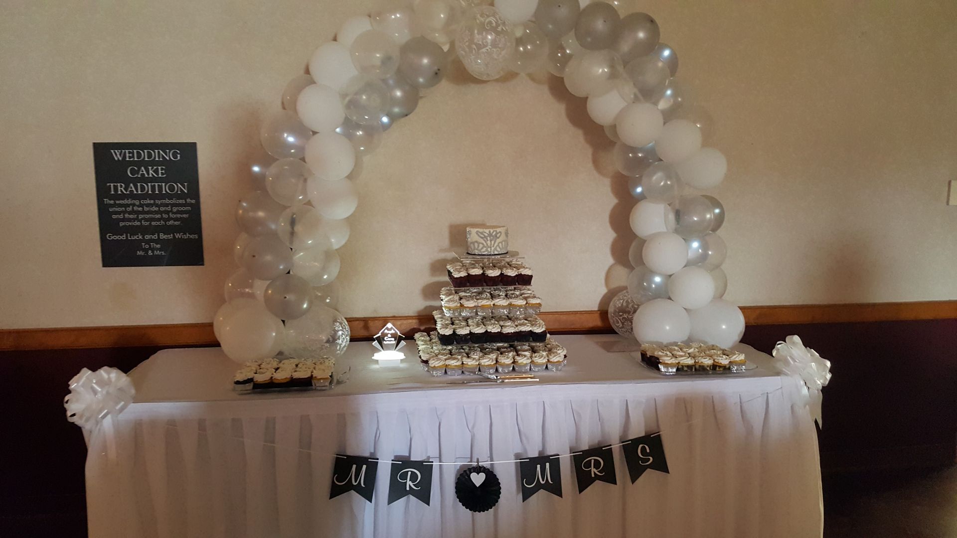 White balloon arch over a decorated dessert table with a tiered cake and black bunting banner for an event