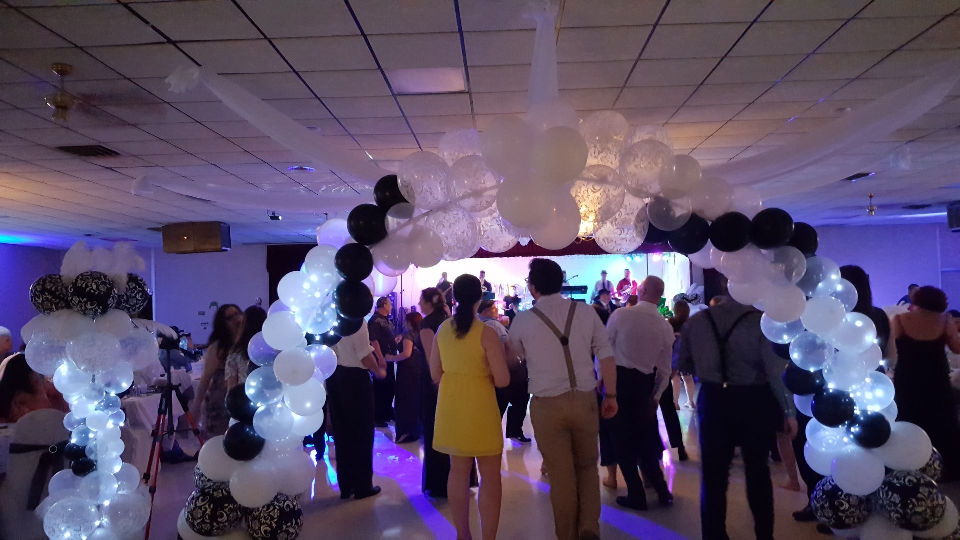 Guests gather under a black-and-white balloon arch at a purple-lit indoor party.