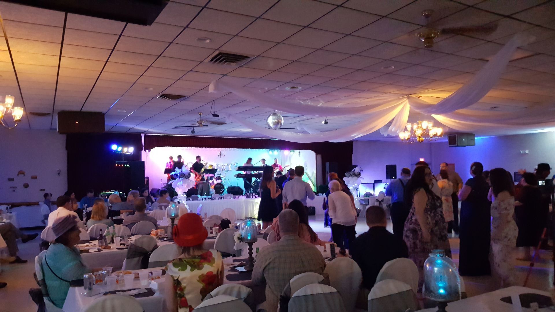 Banquet hall reception with guests seated and dancing near a lit stage under draped ceiling decor
