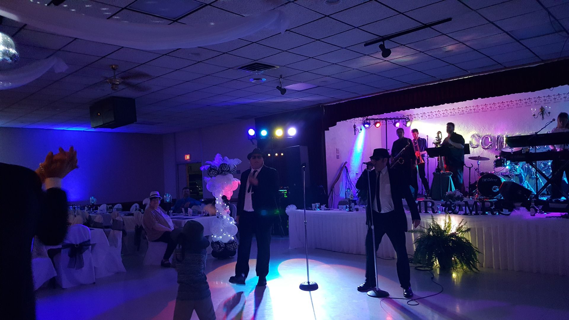 Dimly lit banquet hall with guests dancing near a stage bathed in blue and purple lights.