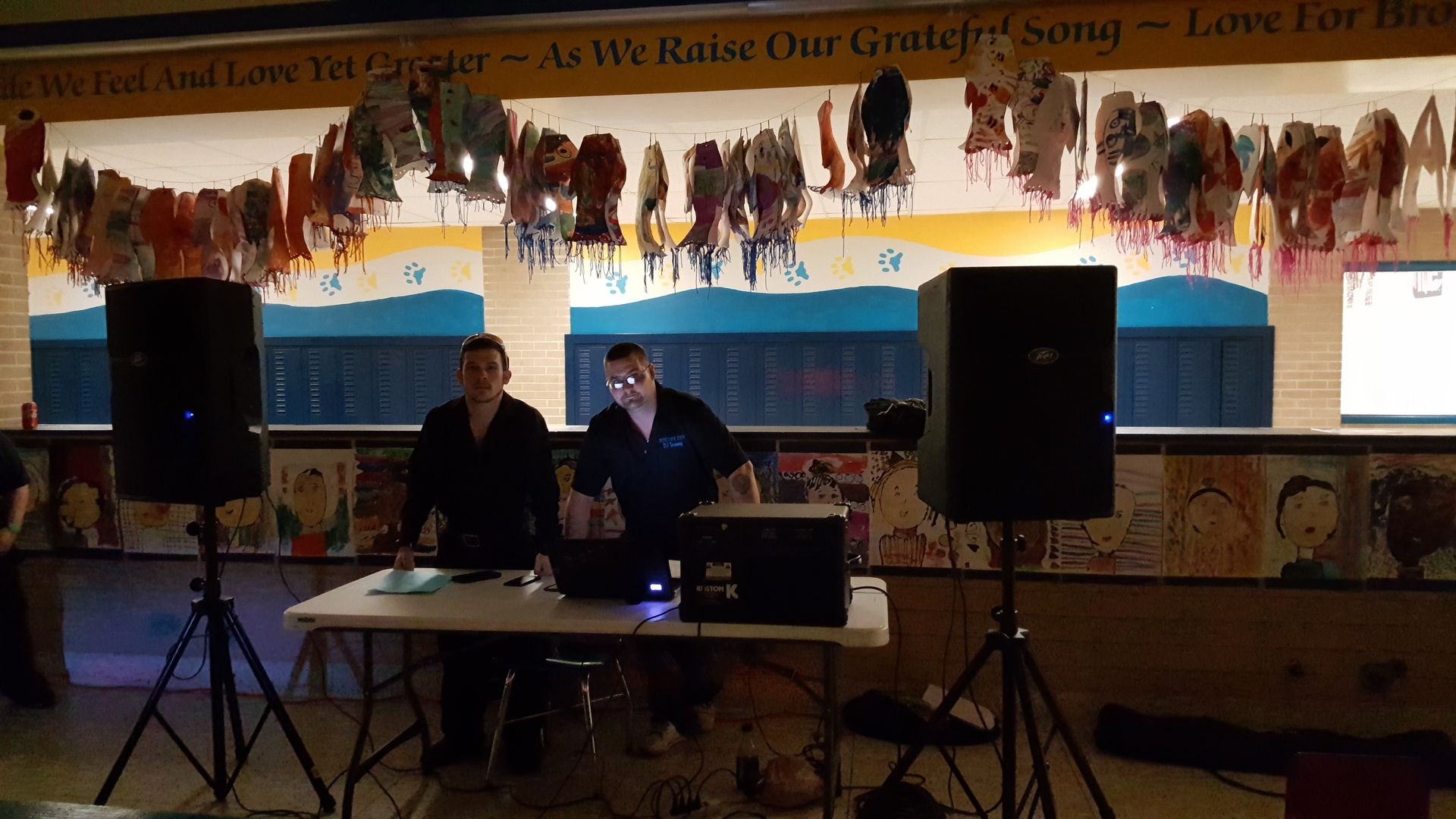 Two people at laptops on a small stage with speakers in a dimly lit venue, under a colorful mural.