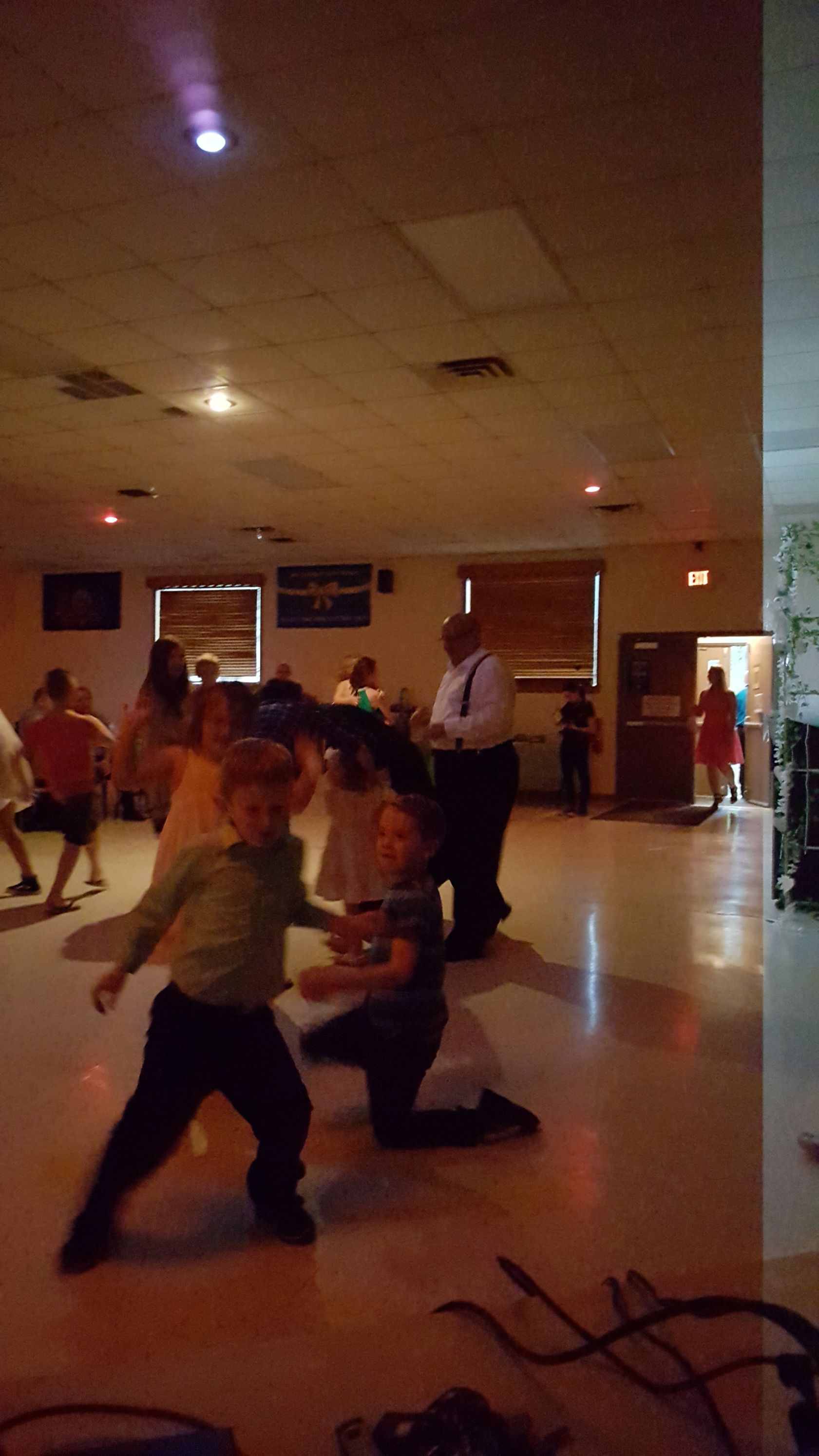 People dancing in a dimly lit hall with a ceiling fan and wall windows