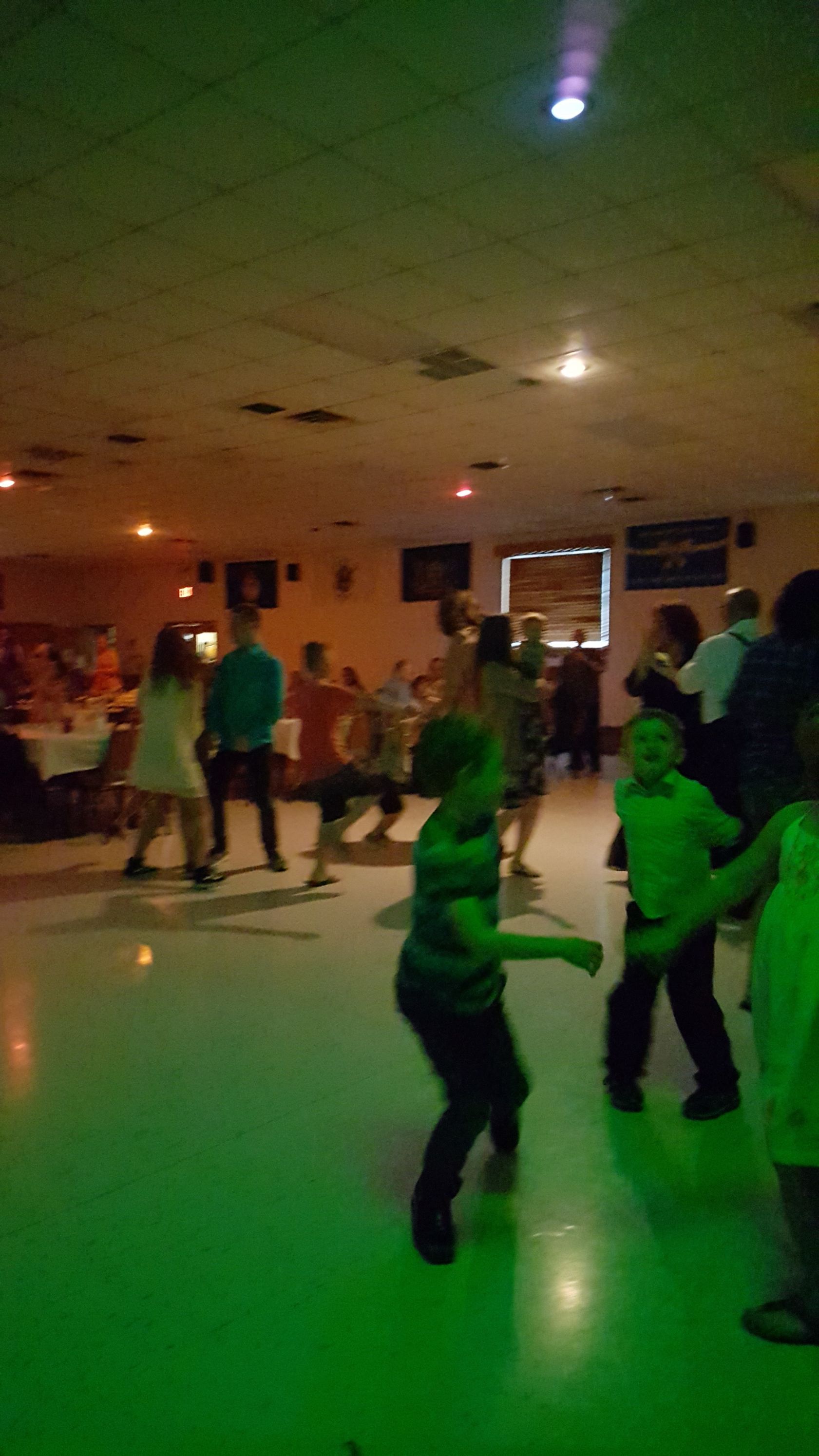 People dancing in a dimly lit room with green floor reflections and ceiling lights.
