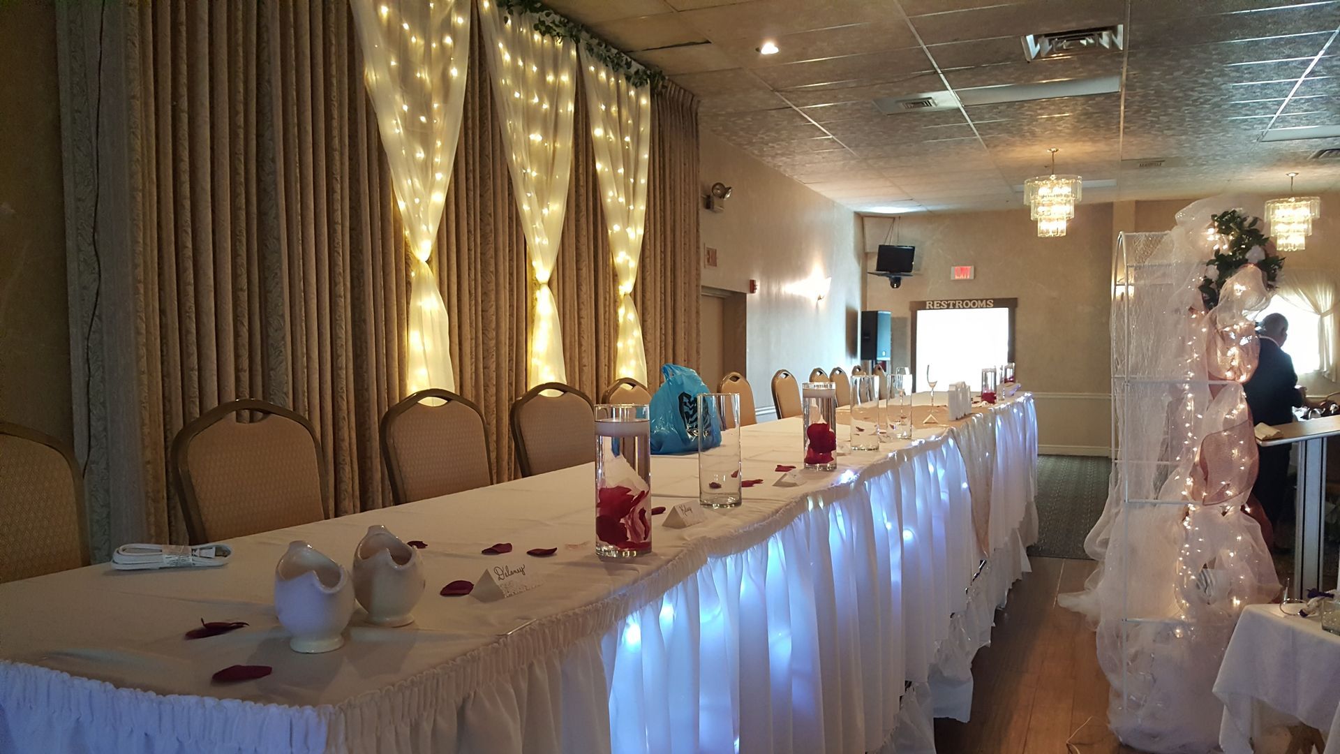 Decorated banquet hall with a long white table, twinkling lights, and a wedding cake display on the right