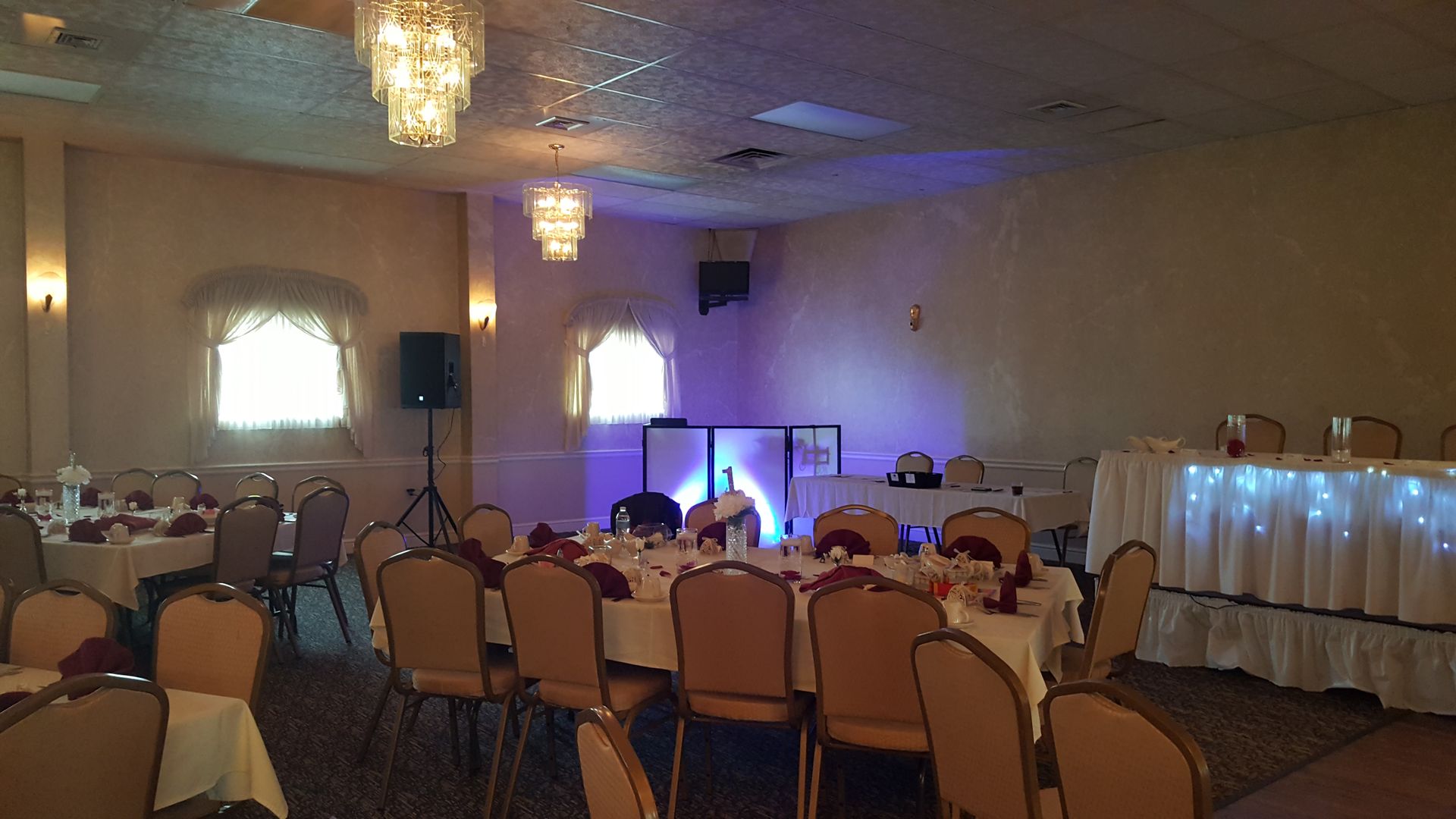 Banquet hall with rows of chairs, chandeliers, and a lit DJ booth near a draped head table