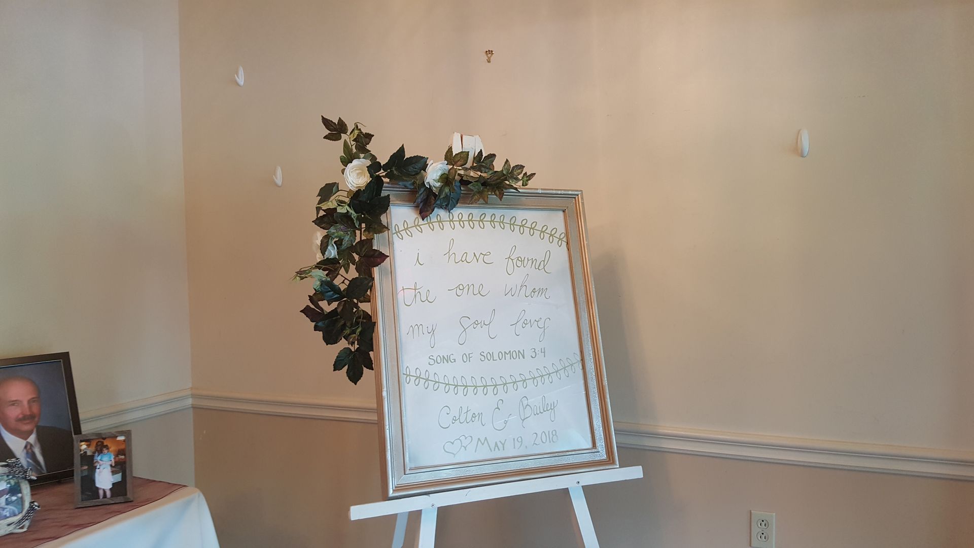 Wedding reception sign on an easel beside a table with framed photos and floral greenery.
