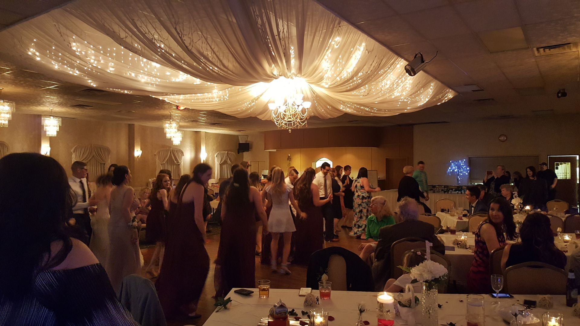 Elegant indoor wedding reception with guests dancing under draped ceiling lights and a chandelier