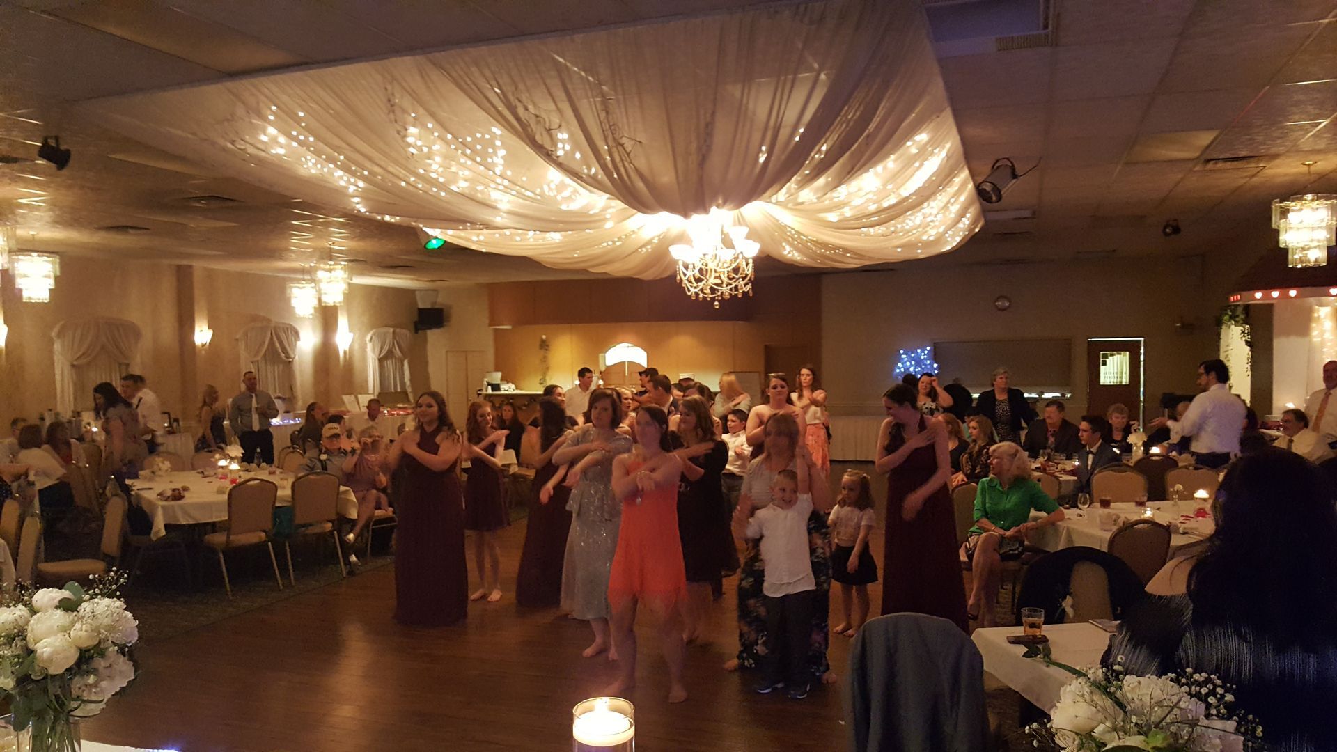 Guests dance at a warmly lit banquet hall with draped ceiling decor and chandeliers.