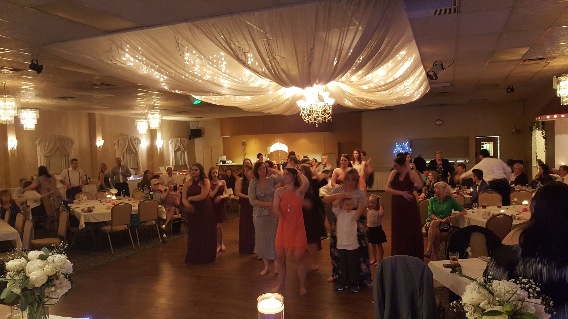 Guests dance in a warmly lit banquet hall with draped ceiling decor and seated tables.