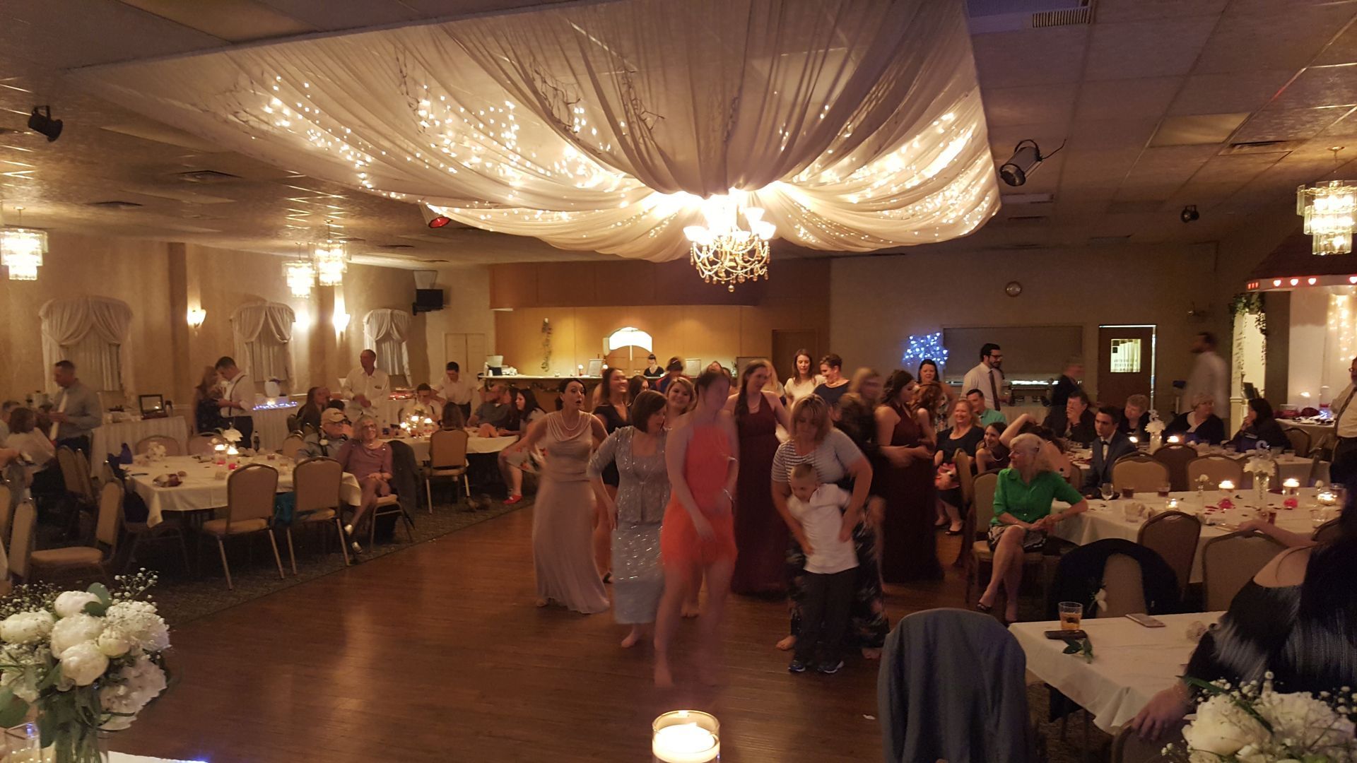 Guests dance at a warmly lit banquet hall reception under draped white fabric and chandeliers.