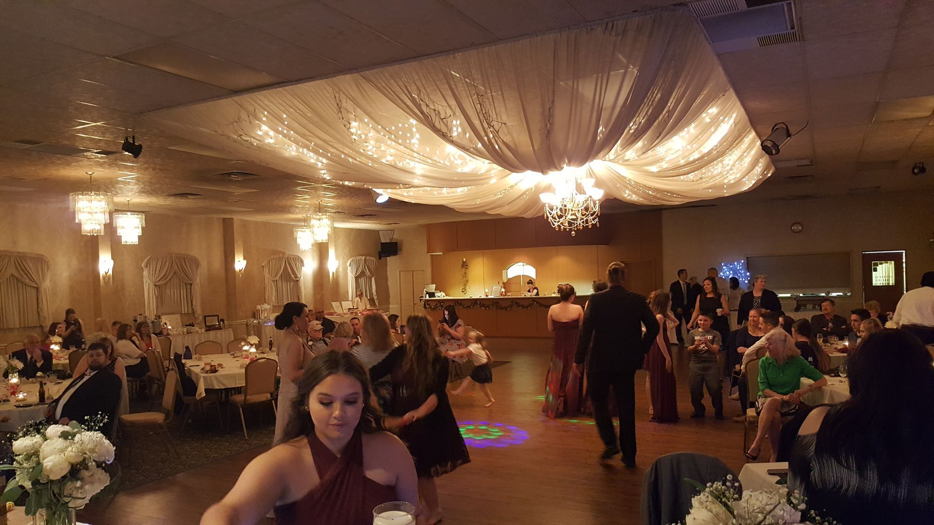 Banquet hall with guests seated at tables under draped ceiling lights, people mingling on the dance floor.