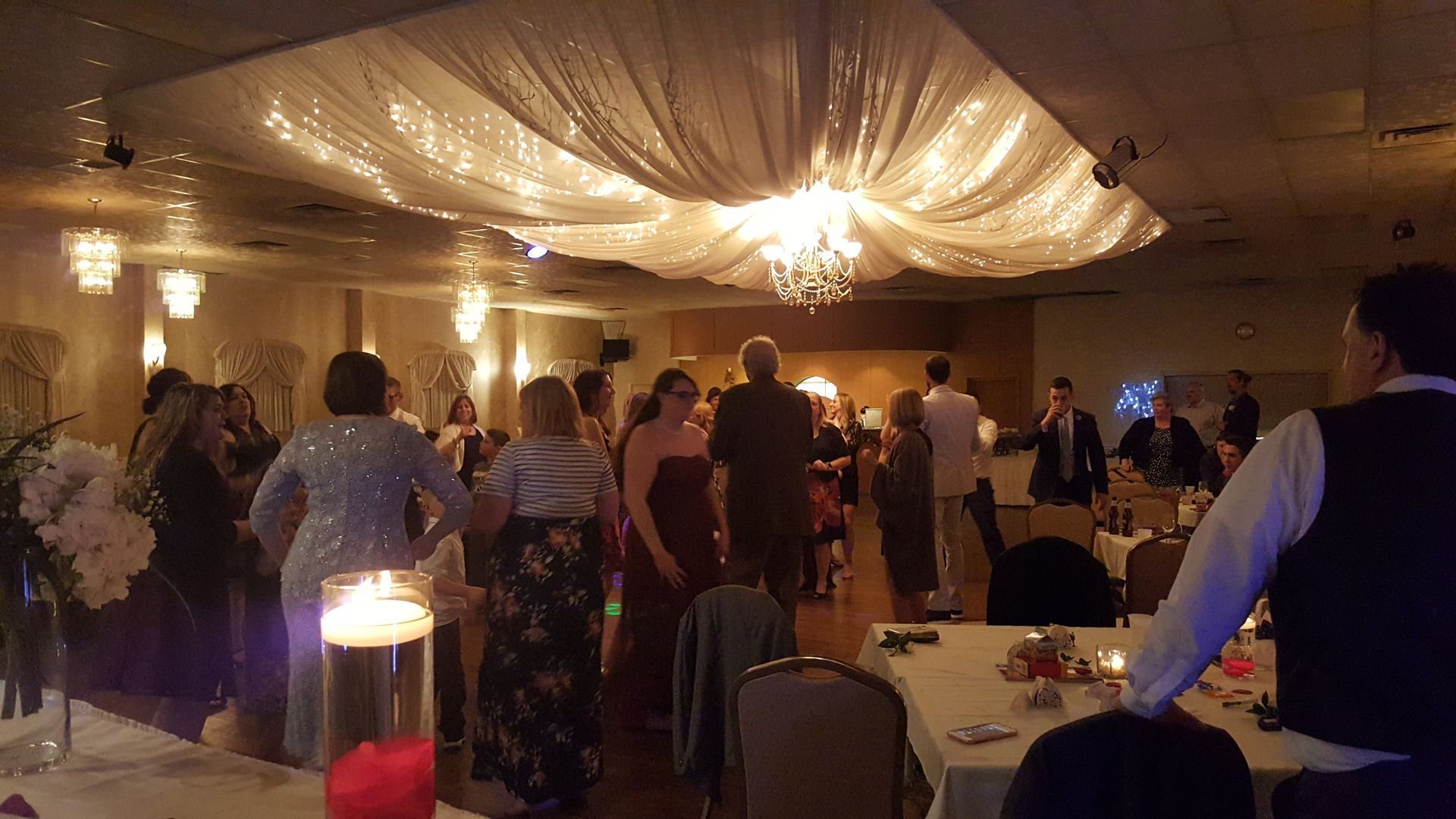 Guests mingle at a warmly lit indoor reception under draped white ceiling fabric and chandeliers.