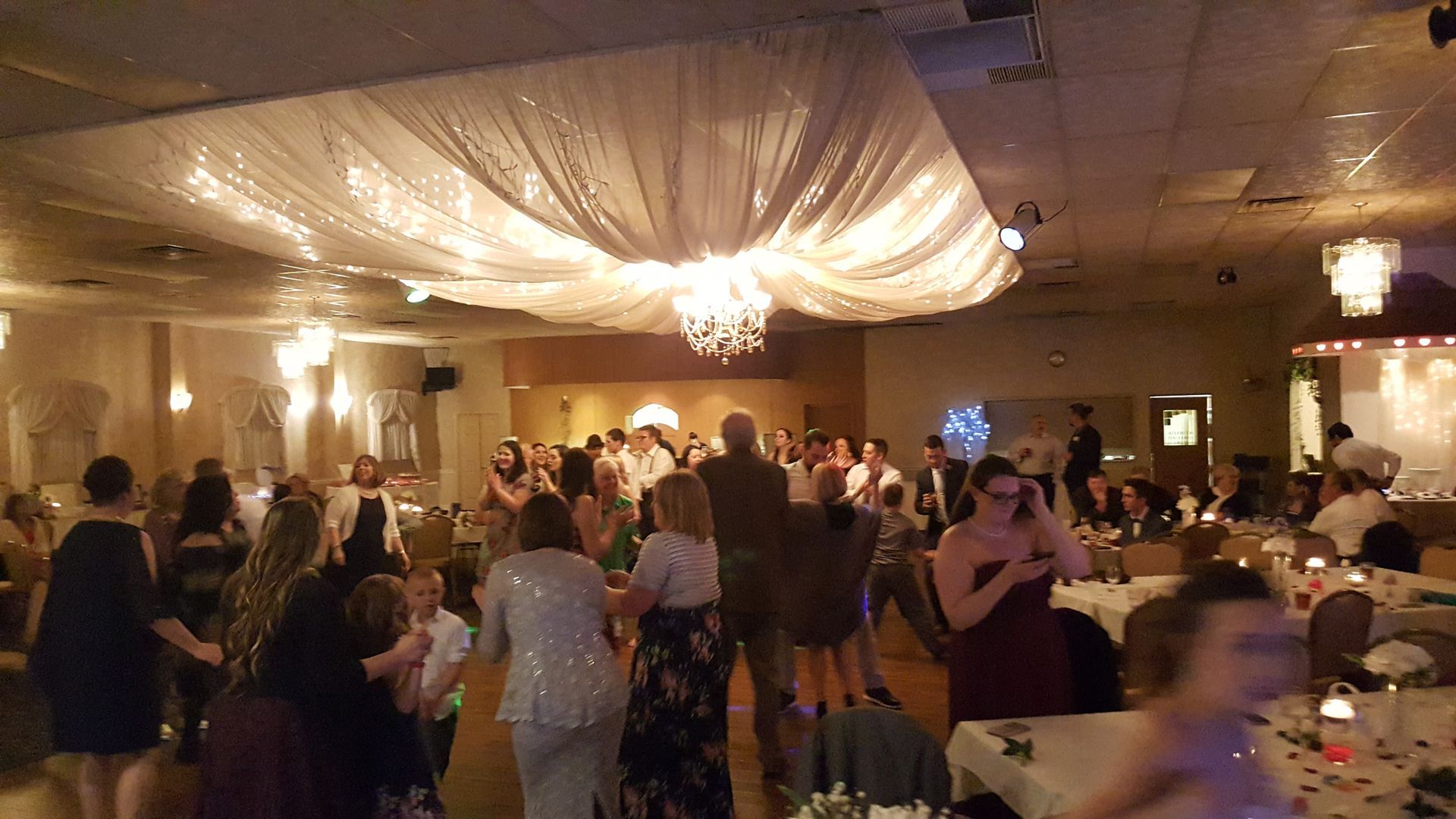 Guests dance and mingle in a warmly lit banquet hall with draped ceiling fabric and round tables.