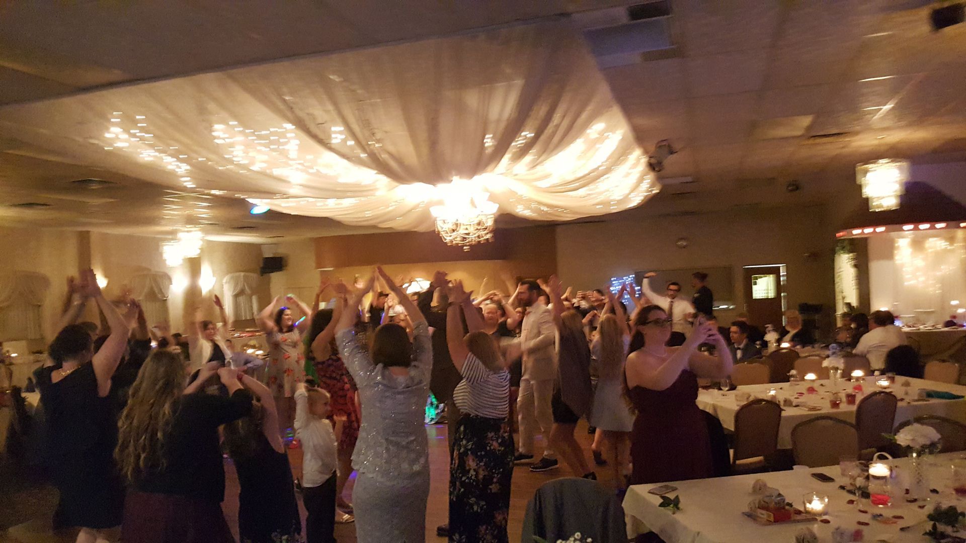 People dancing with raised arms at a lively banquet hall reception under warm lighting.