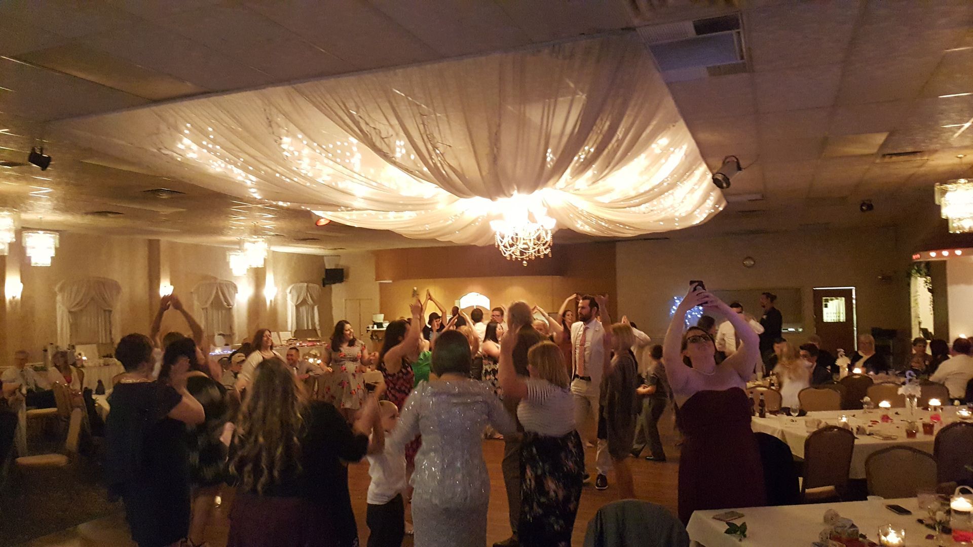 Guests dancing at a warmly lit banquet hall reception under draped ceiling lights.