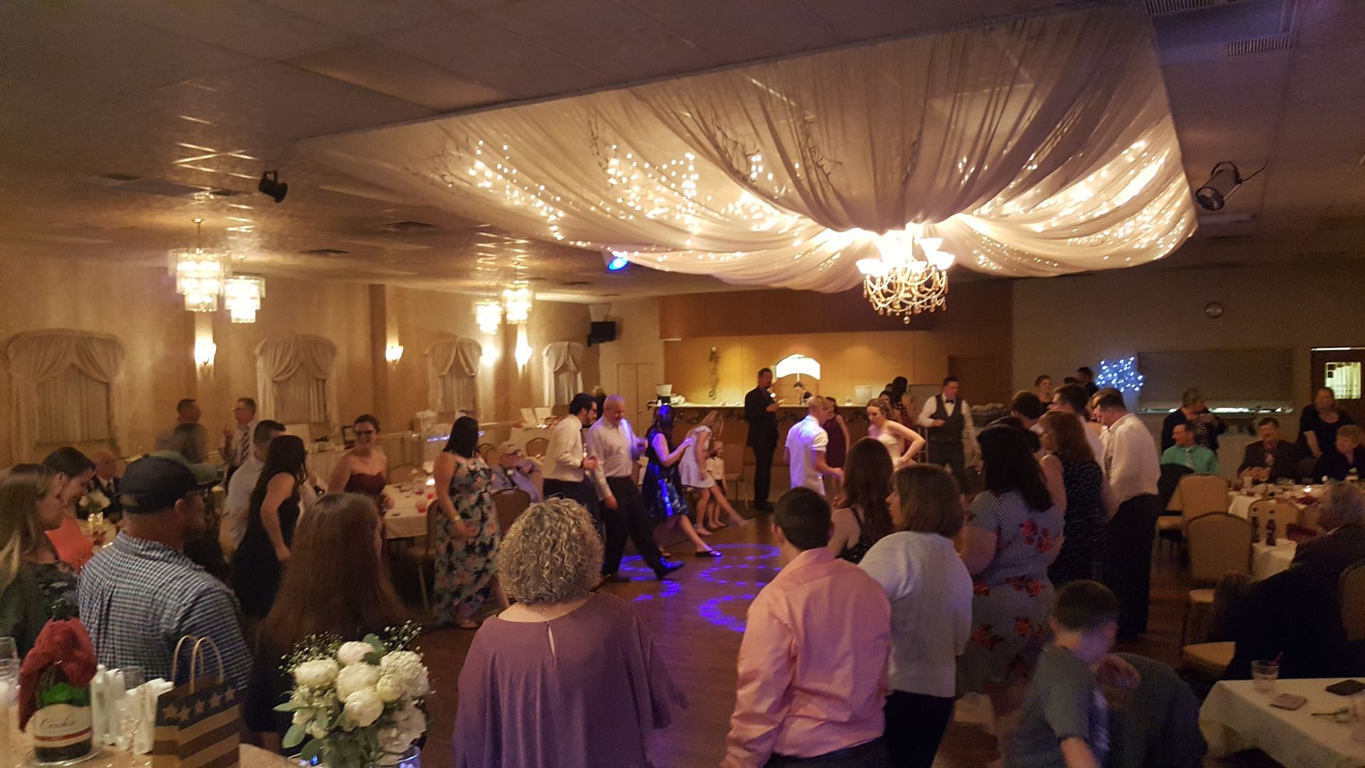 Guests dancing at a warmly lit reception under draped white ceiling fabric and chandeliers