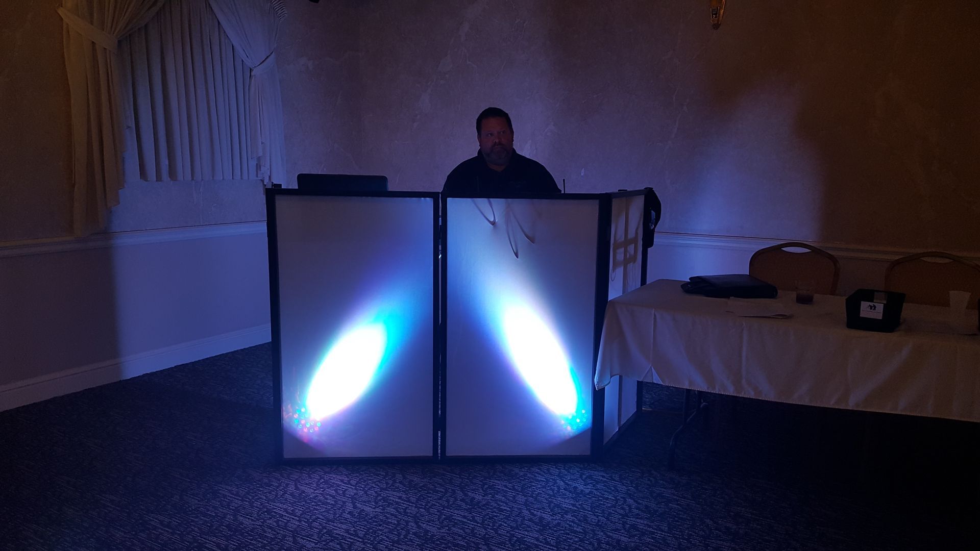 Person behind two glowing blue-lit panels in a dim room, with tables and equipment nearby.
