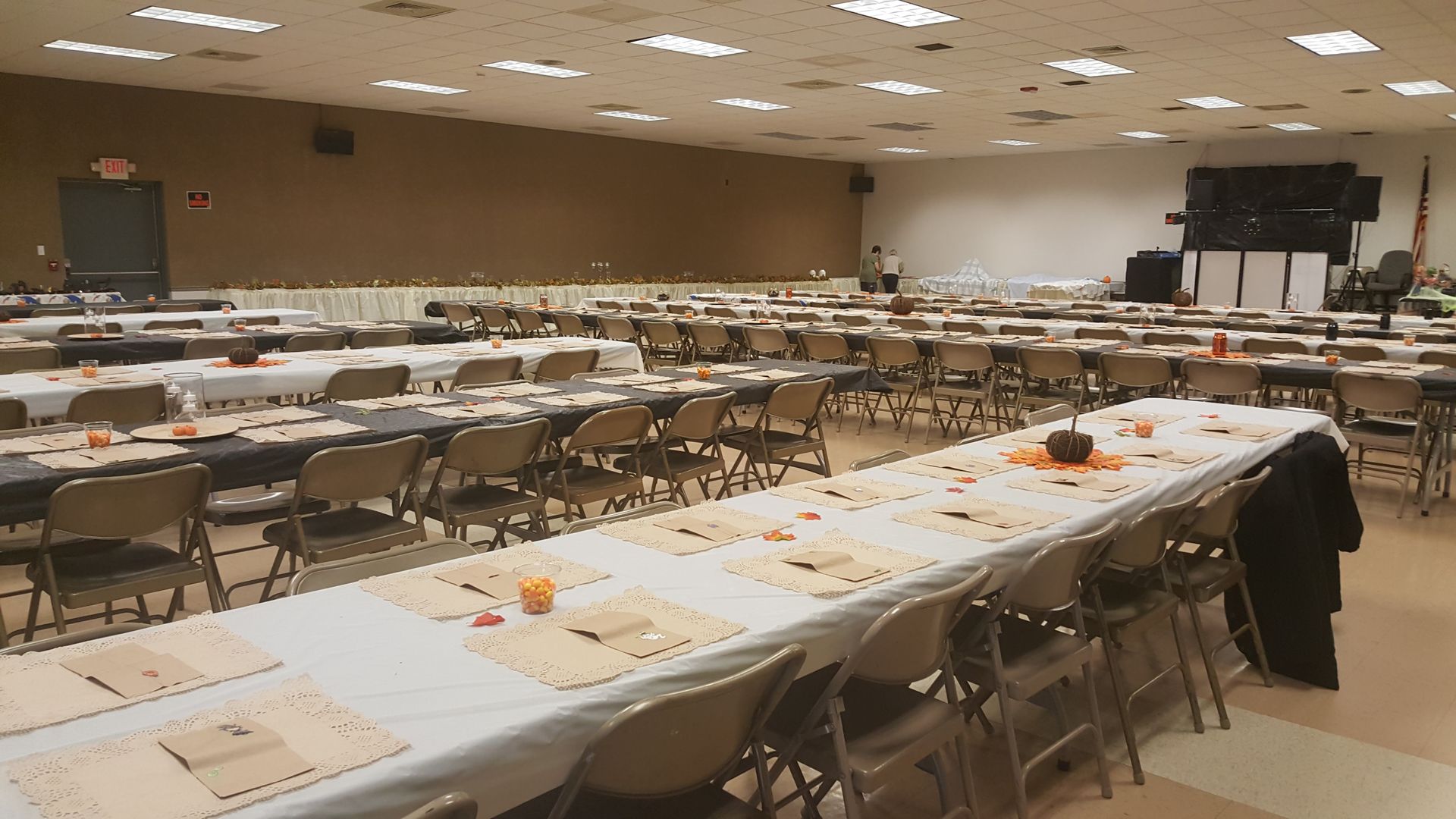 Large banquet hall with long tables, folding chairs, and scattered papers, set up for an event or meeting