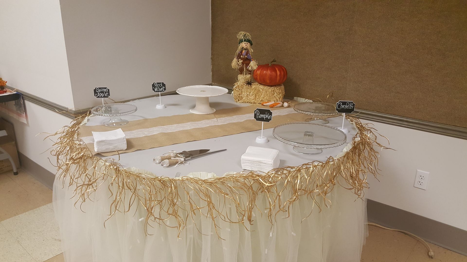 Decorated buffet table with white cloth, gold fringe, plates, utensils, and centerpieces in a banquet room