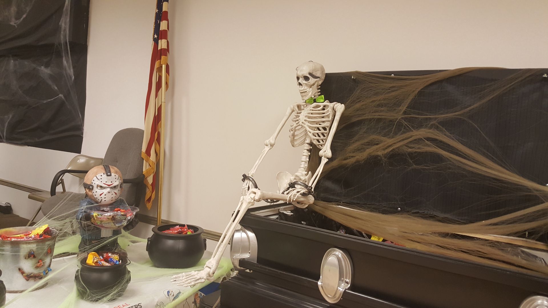 Skeleton decorations beside black Halloween webbing on a bench, with bowls of candy and an American flag nearby