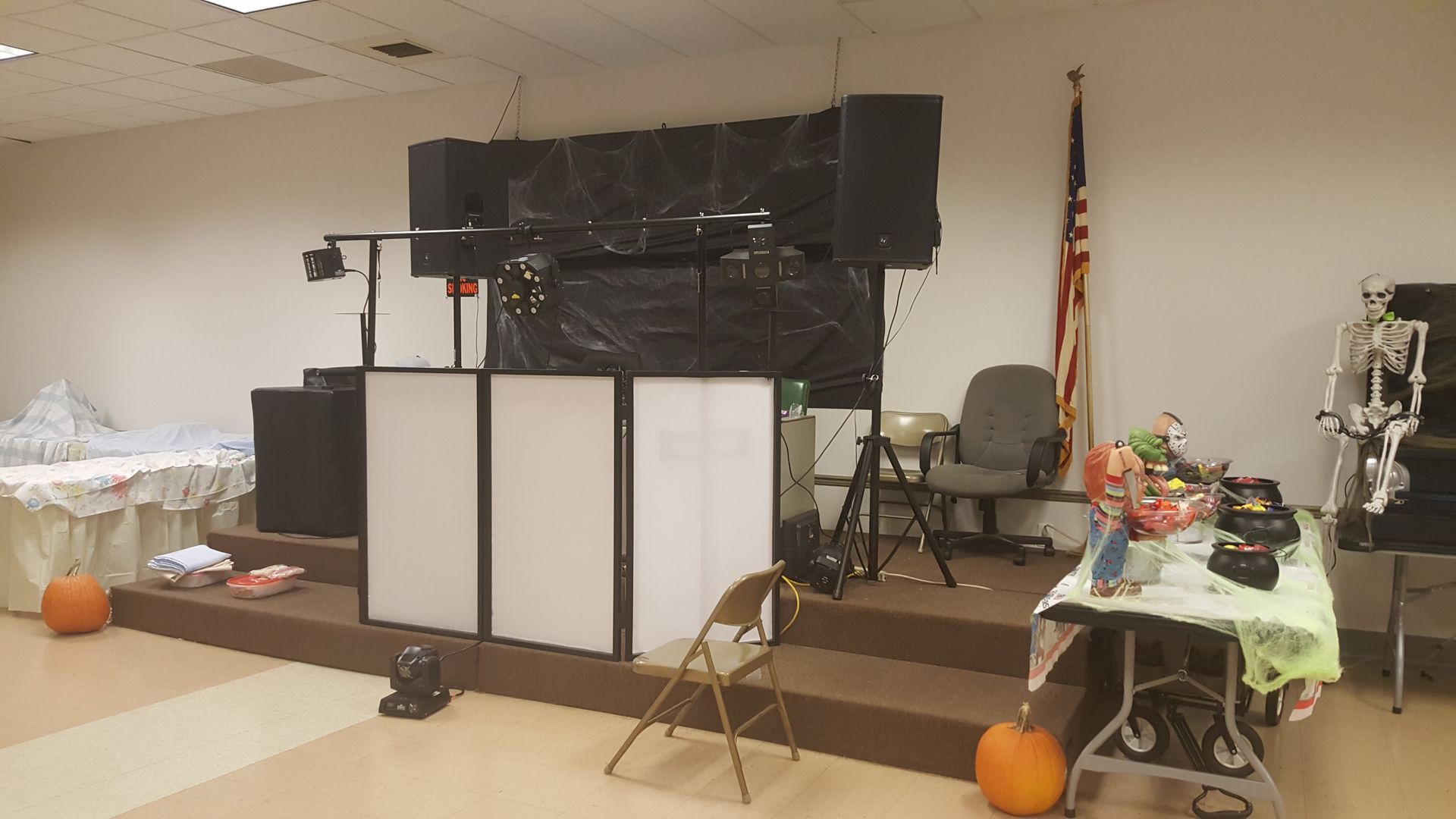 Indoor event setup with large speakers, white DJ booth, tables, and Halloween pumpkins in a room