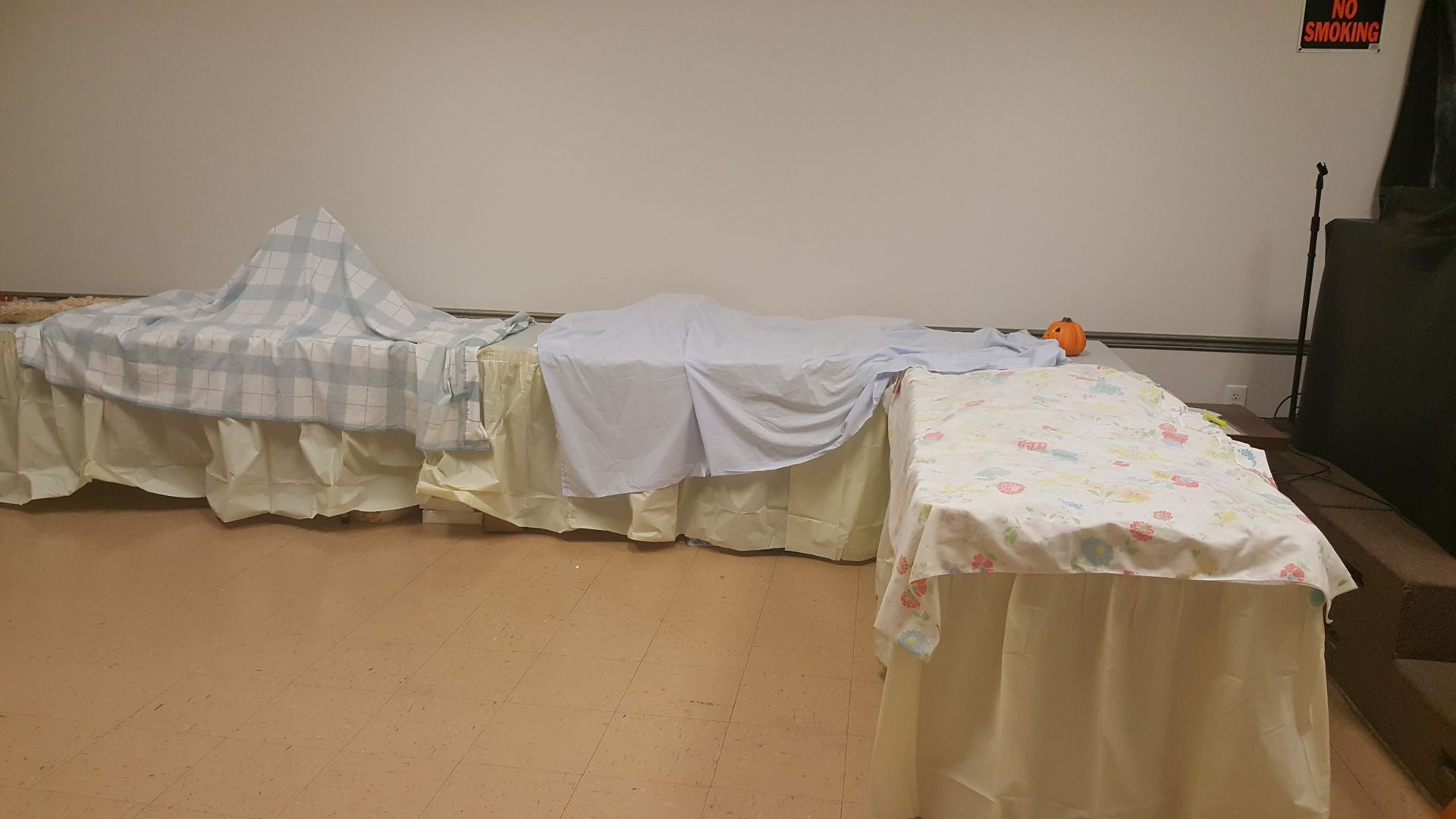 Two covered beds or tables in a plain room, one draped in white and one in a checkered cloth.
