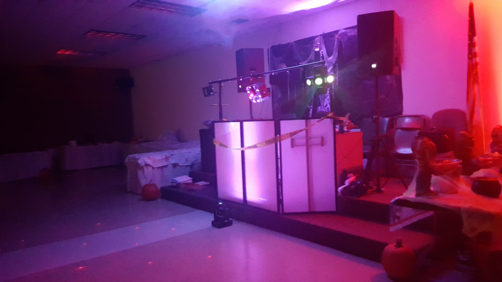 Purple-lit room with white DJ booths and equipment, a small stage, and tables along the right side.
