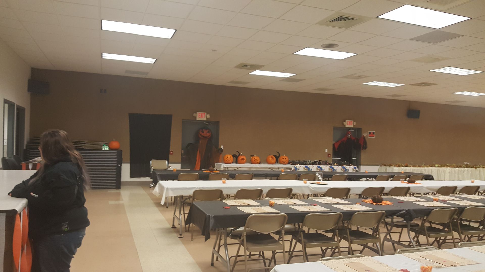 Indoor community hall with long tables and chairs set up for an event, with a few people near the back wall.