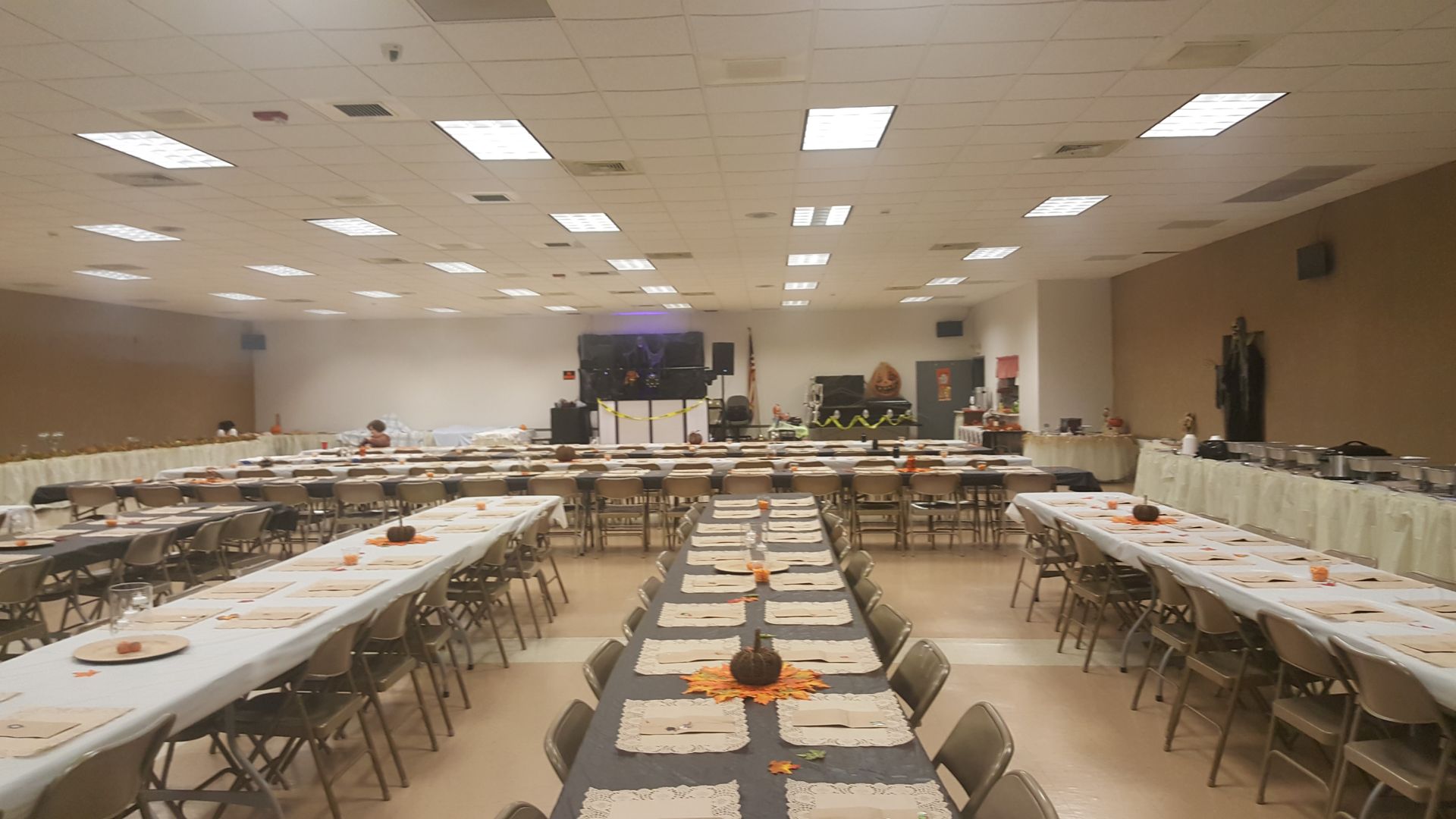 Large banquet hall with long tables, chairs, and a stage at the far end under bright ceiling lights.