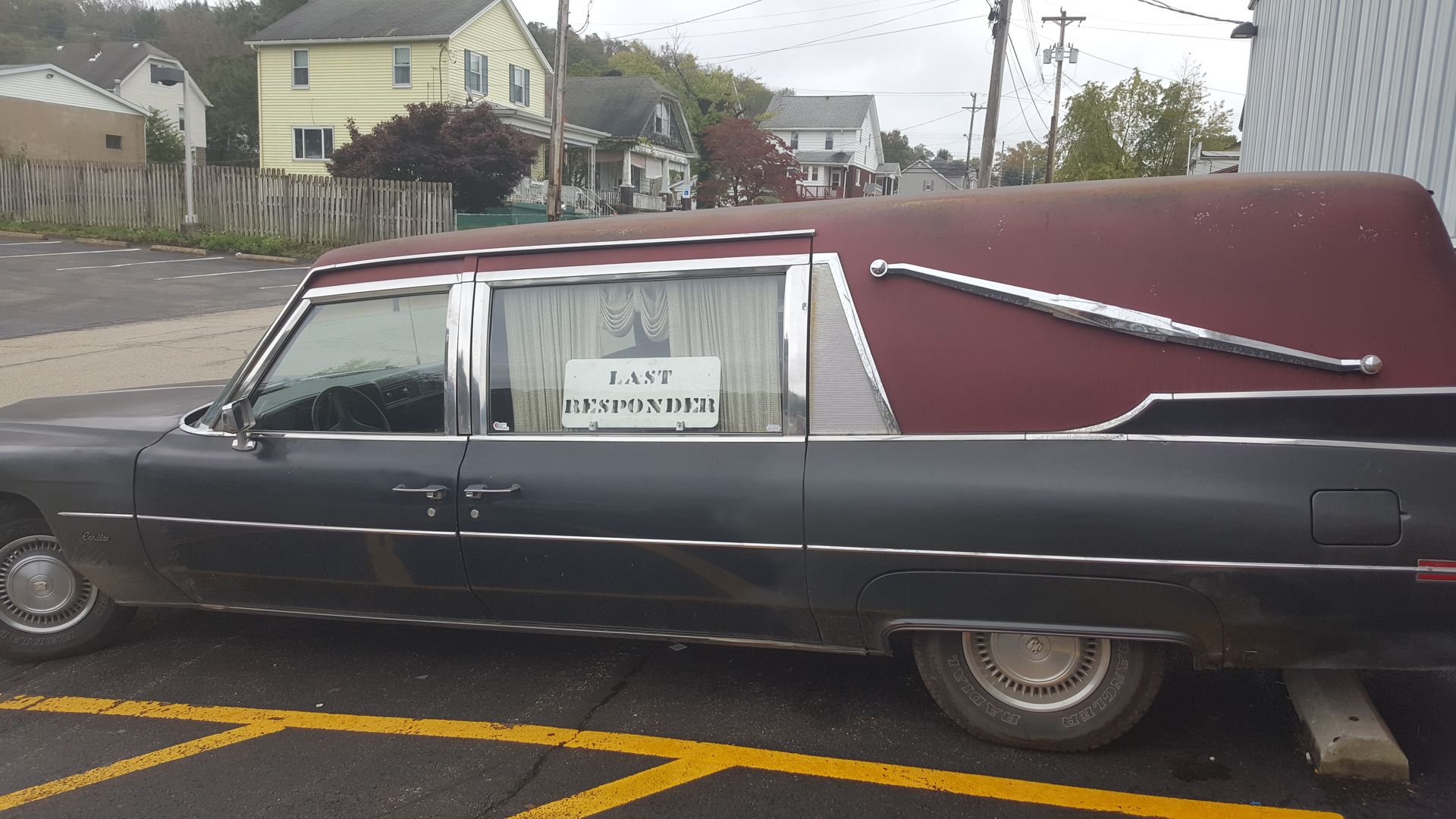 Dark gray vintage hearse parked on a street with a sale sign in the window.