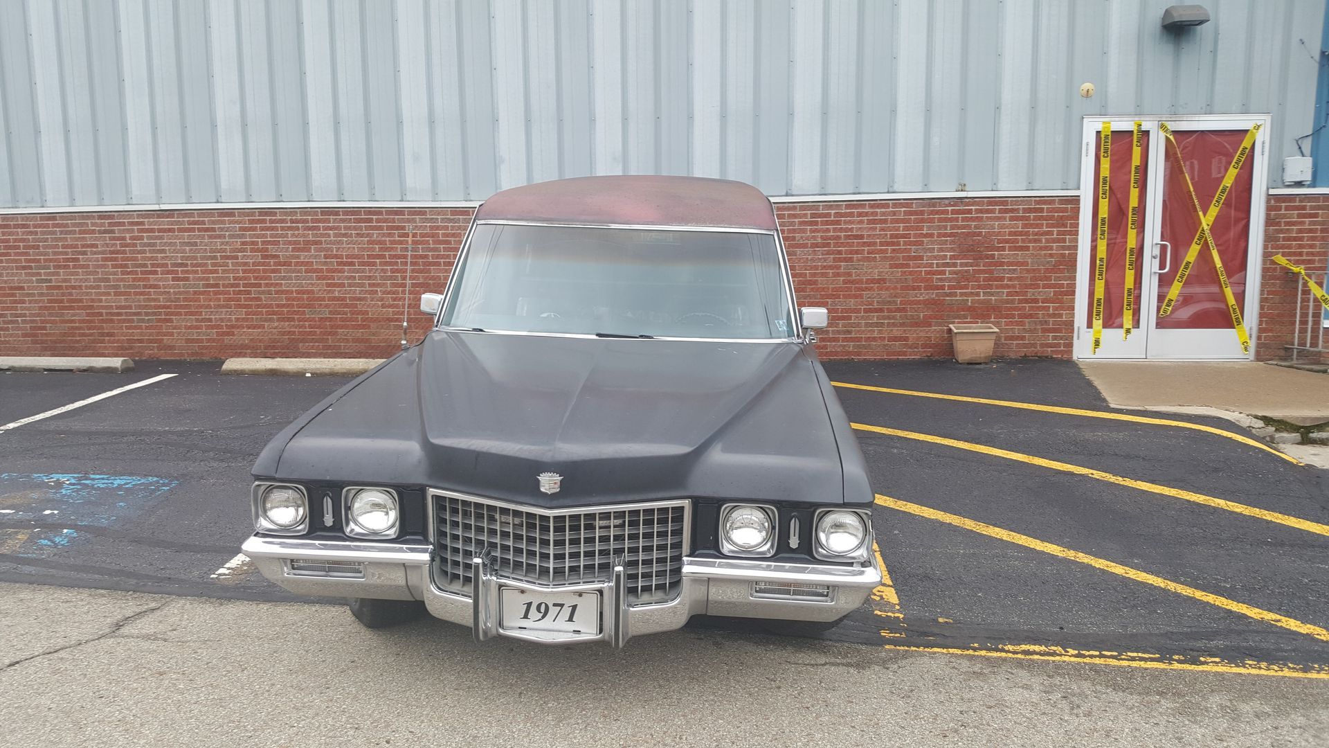 Front view of a black classic car parked outside a building with red brick and gray siding.