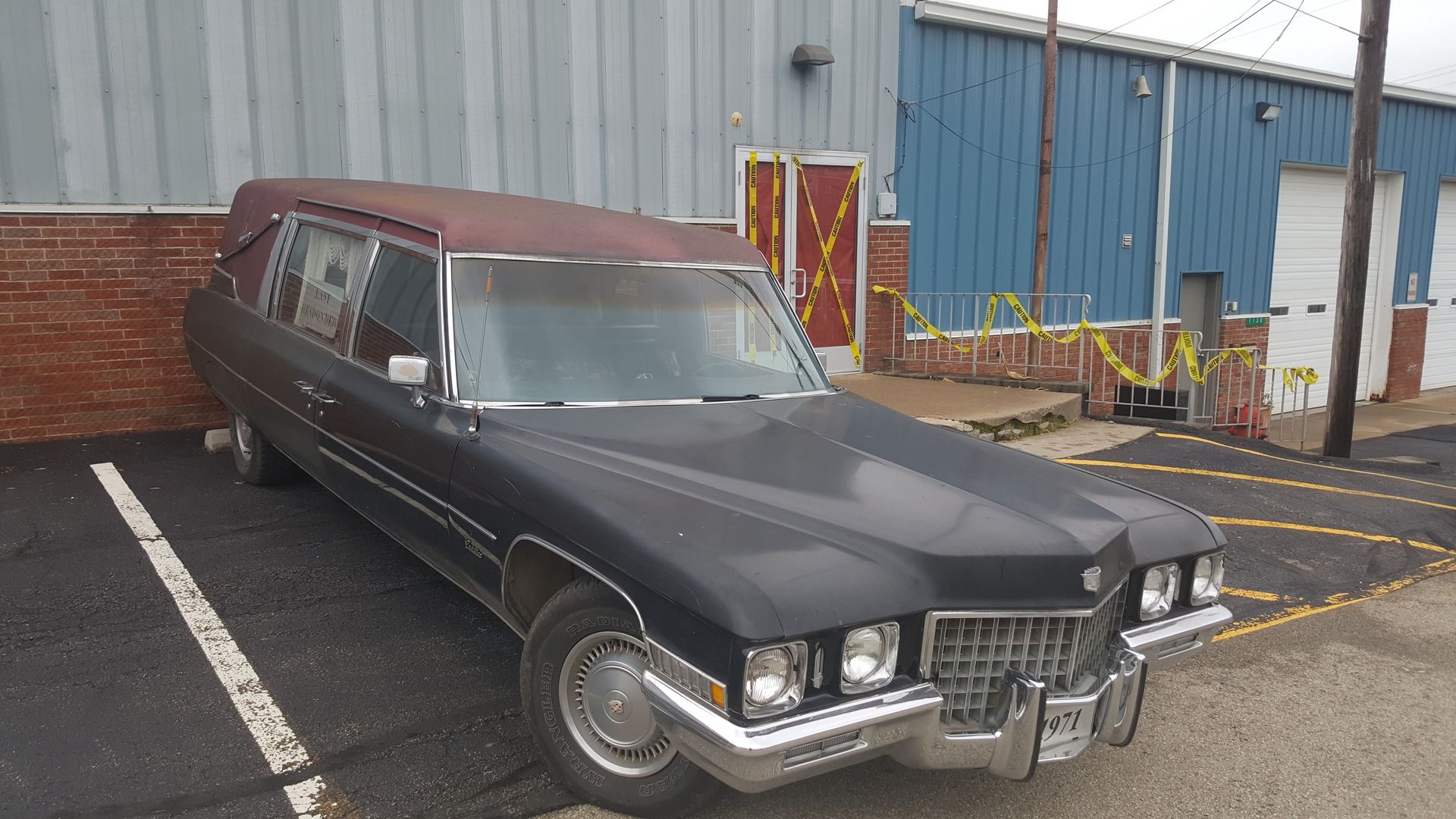 Black classic hearse parked in a lot beside an industrial building.