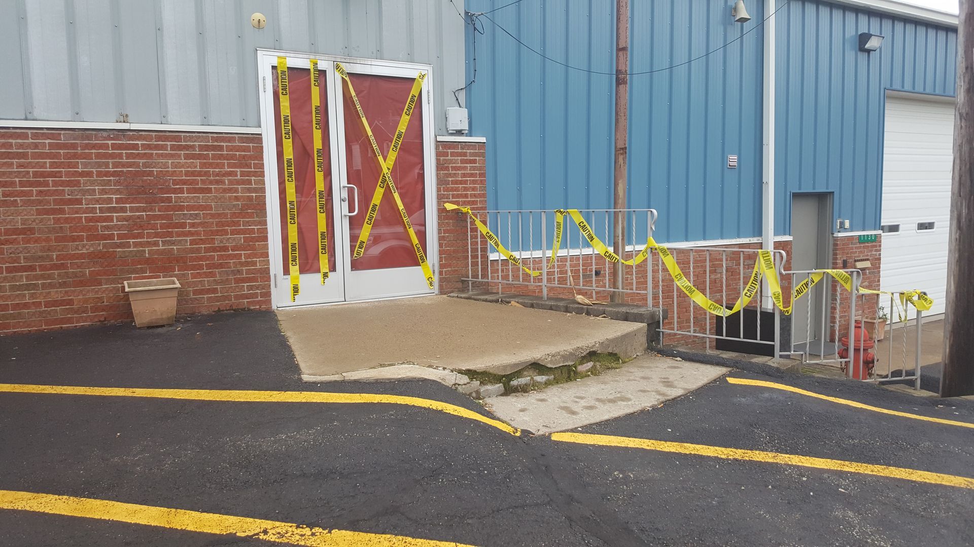Building entrance blocked off with yellow caution tape and boarded red doors beside a ramp.
