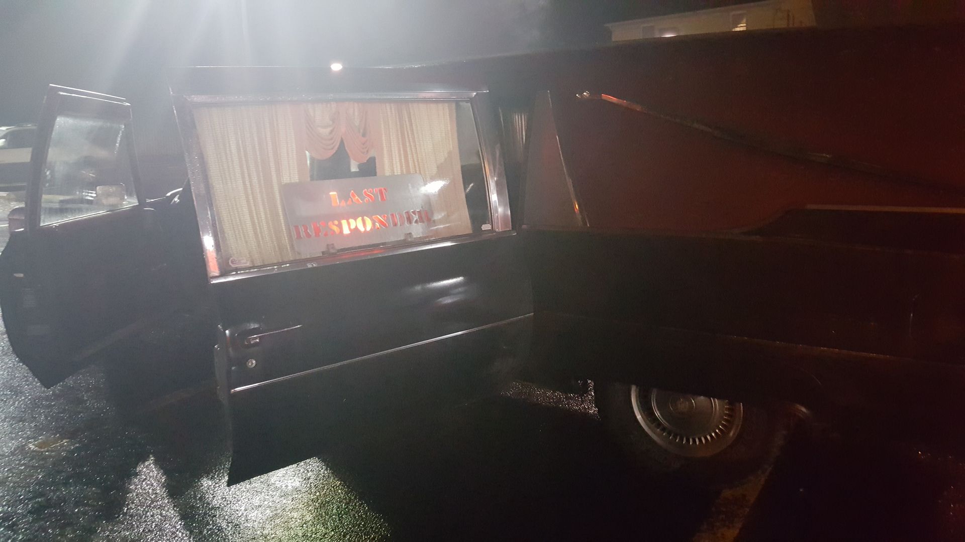 Dark glossy vehicle with an open side door revealing lit interior at night on a wet street