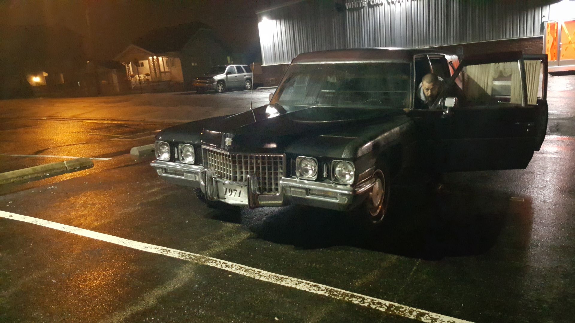 Black vintage sedan parked at night in a wet lot, with a person visible in the open rear door.