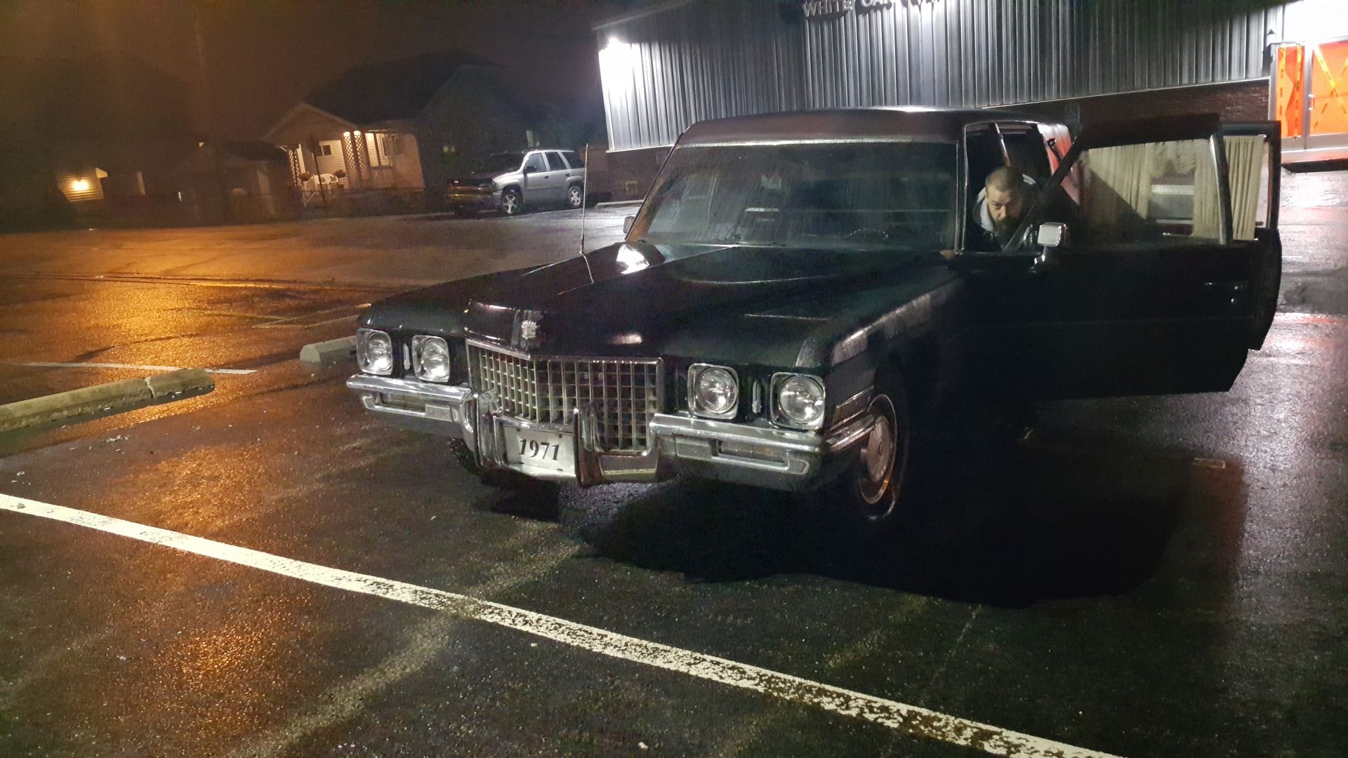 Black vintage car parked on a wet street at night with the driver’s door open.