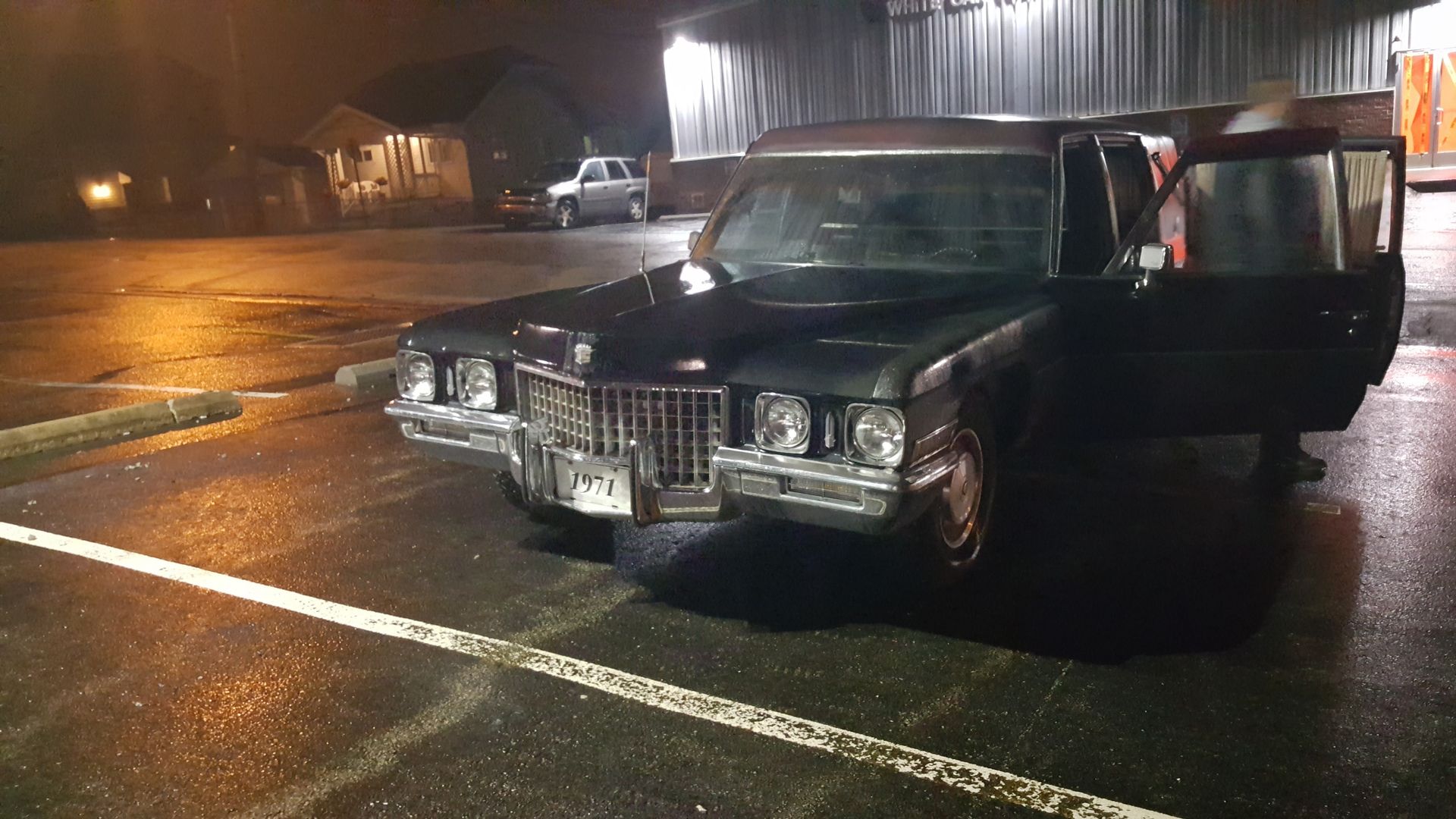 Black vintage car with doors open, parked on a wet lot at night under warehouse lights.