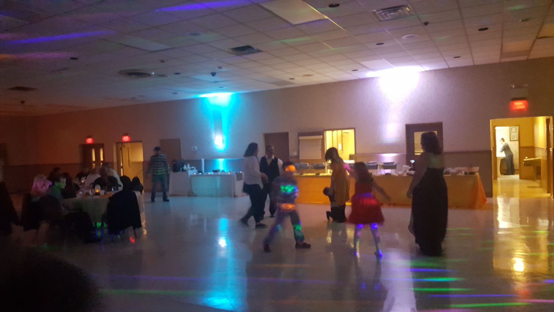 People dancing in a dimly lit banquet hall with colorful lights reflecting on the floor.