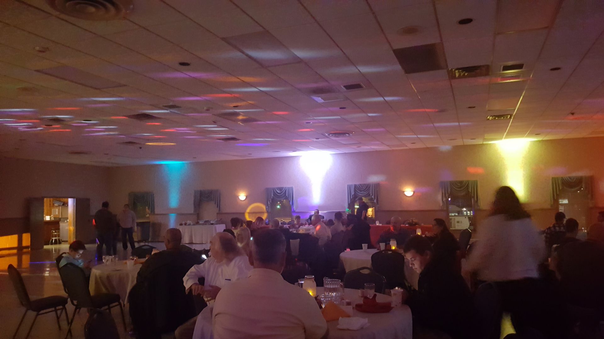 Dim banquet hall with people seated at round tables under colored uplights during an event