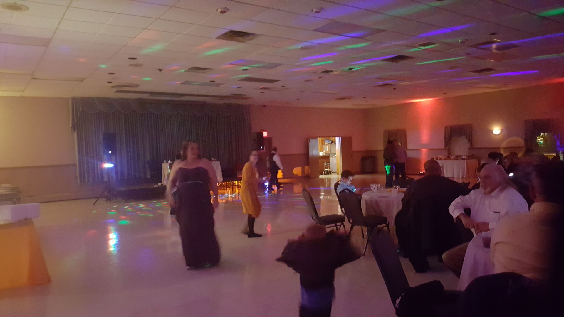 People dancing in a dimly lit banquet hall with colorful ceiling lights.