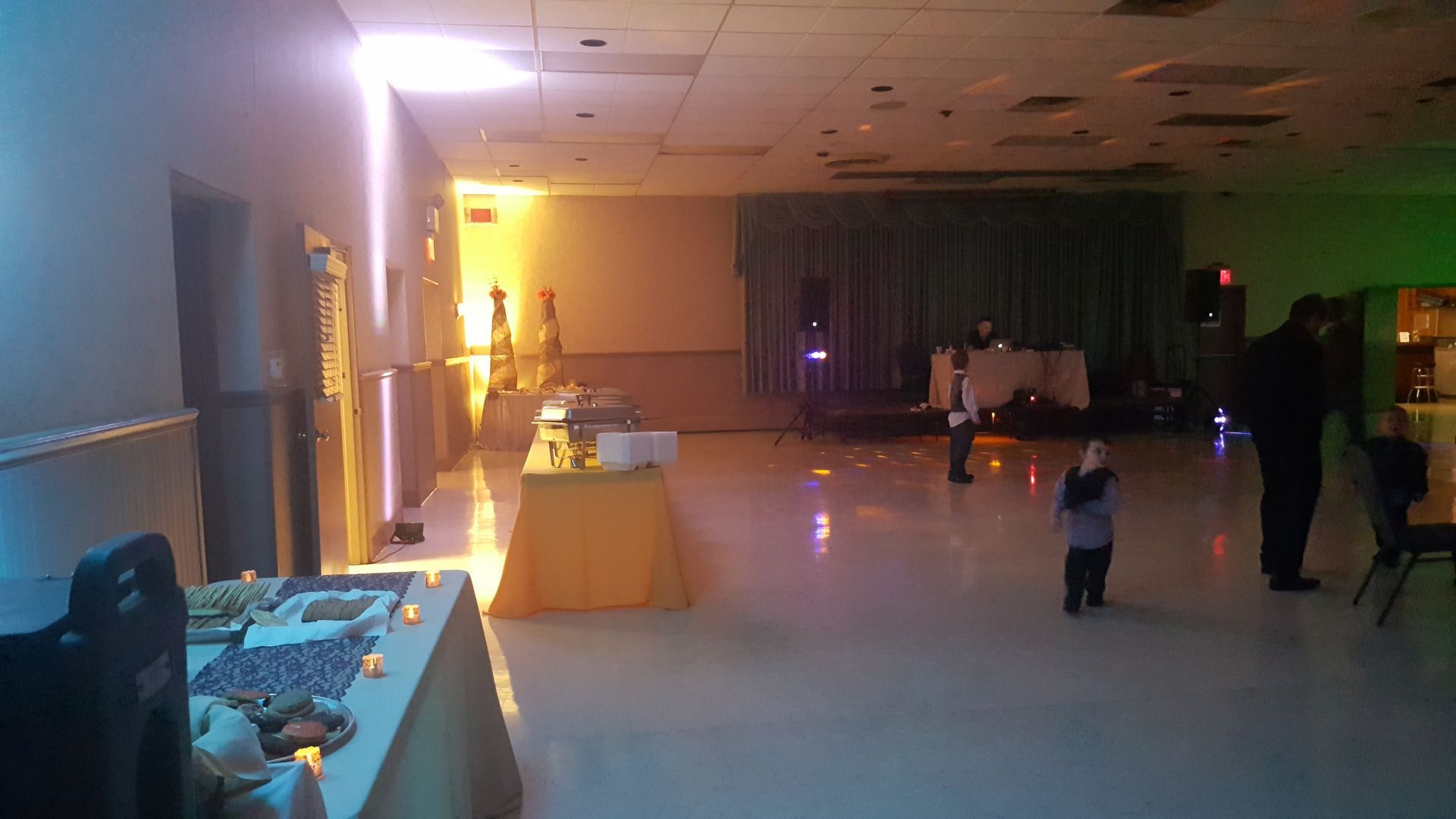 Dim banquet hall with a few people dancing, colored lights, and tables along the walls