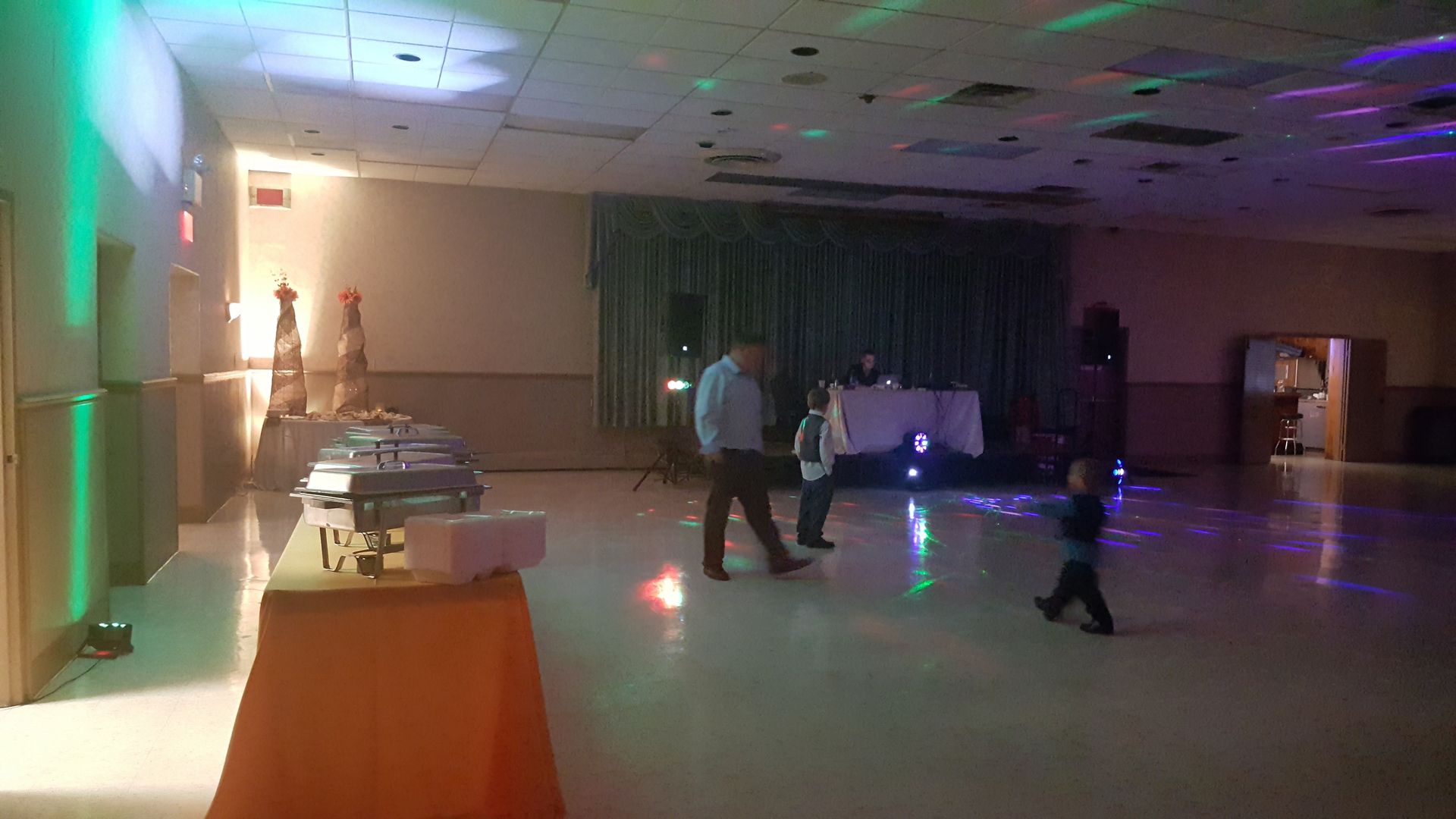 Dimly lit banquet hall with a few people standing near a buffet table and colored lights on the floor