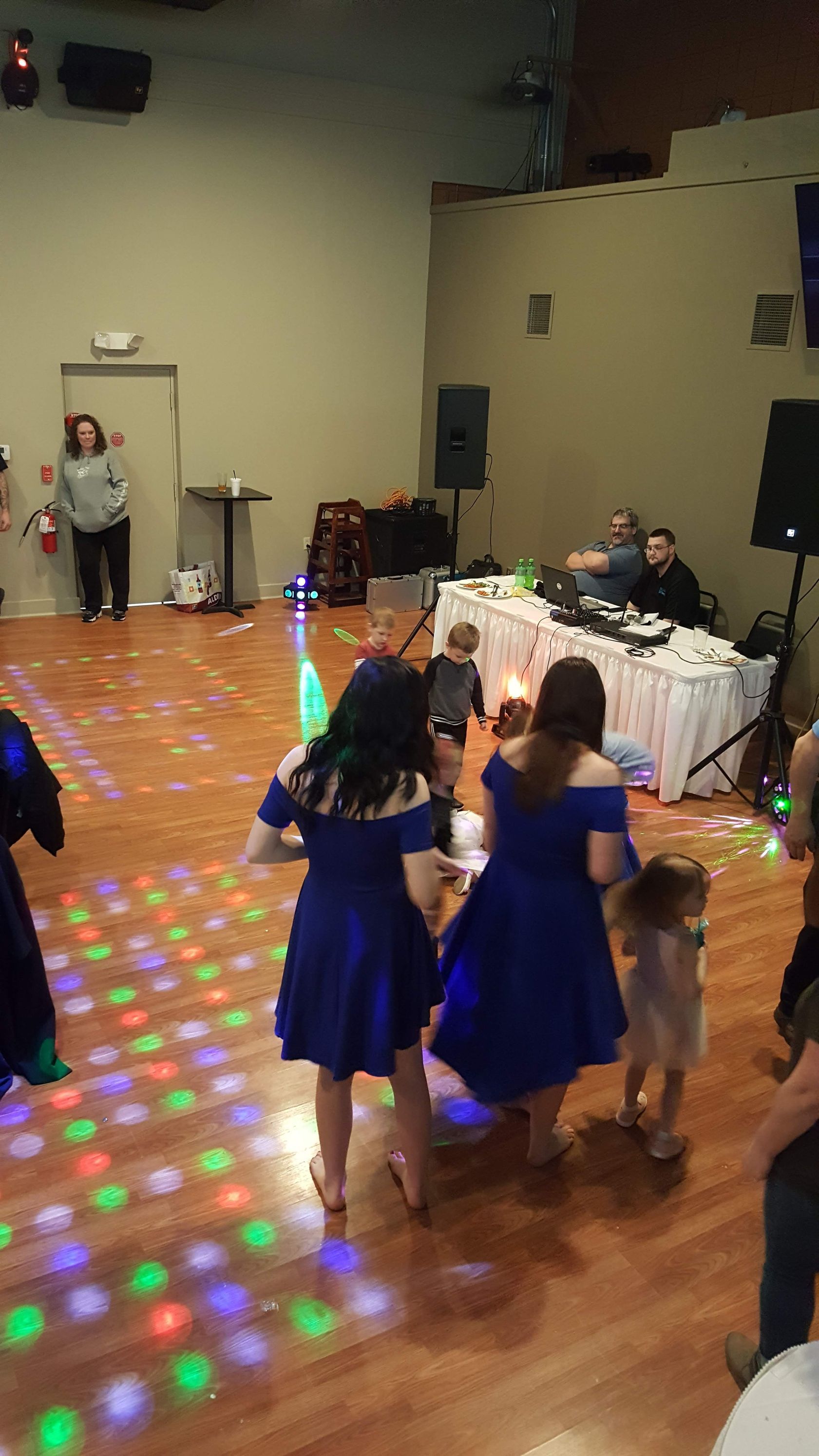 Children in blue dresses dancing on a lit floor at an indoor event with DJ equipment in the background
