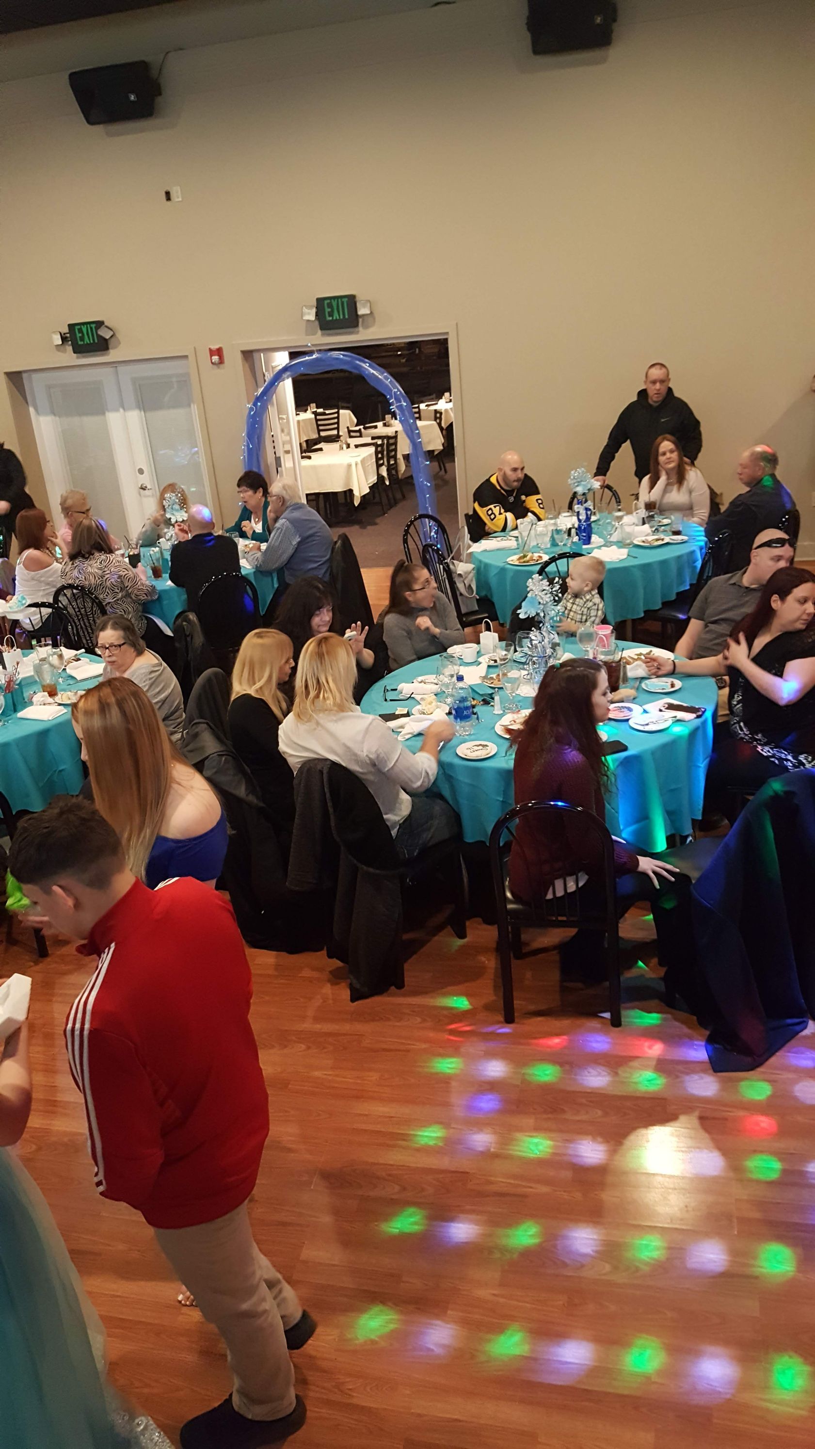 Banquet hall with guests seated at round tables, blue tablecloths, and colorful floor lights on the dance floor