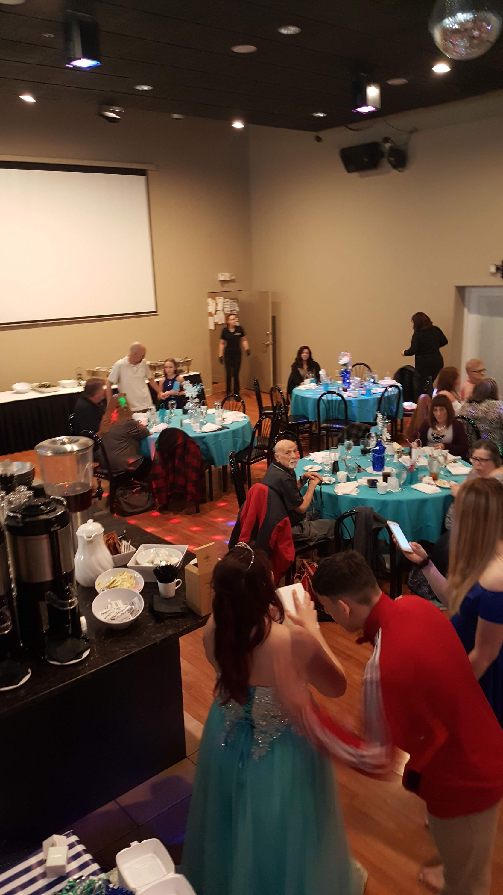 People gathered around decorated tables at a festive indoor event with a projection screen and ceiling lights.