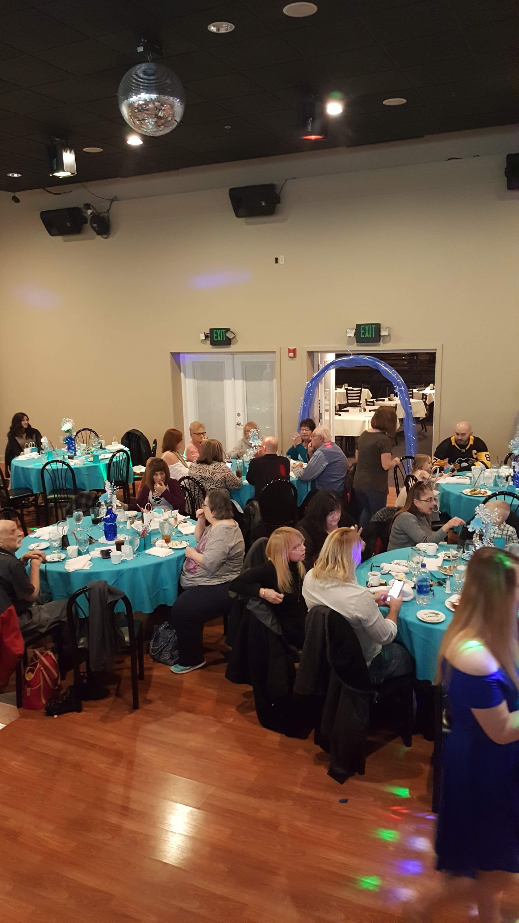 Guests seated at teal-covered tables in a banquet hall, watching an event near an arched backdrop.