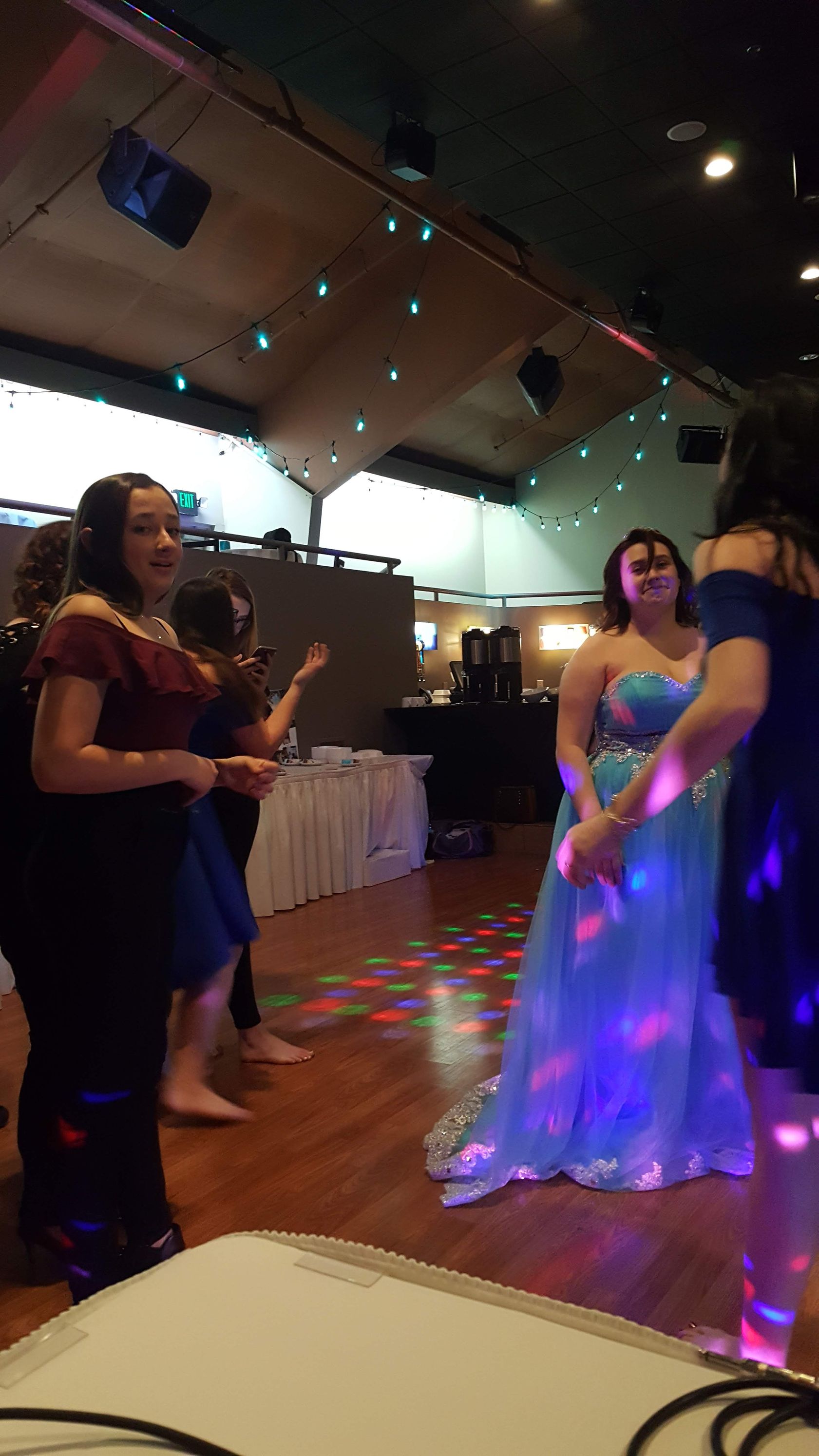 People dancing at a dimly lit party, with colorful lights reflecting on the floor and a woman in a white dress.